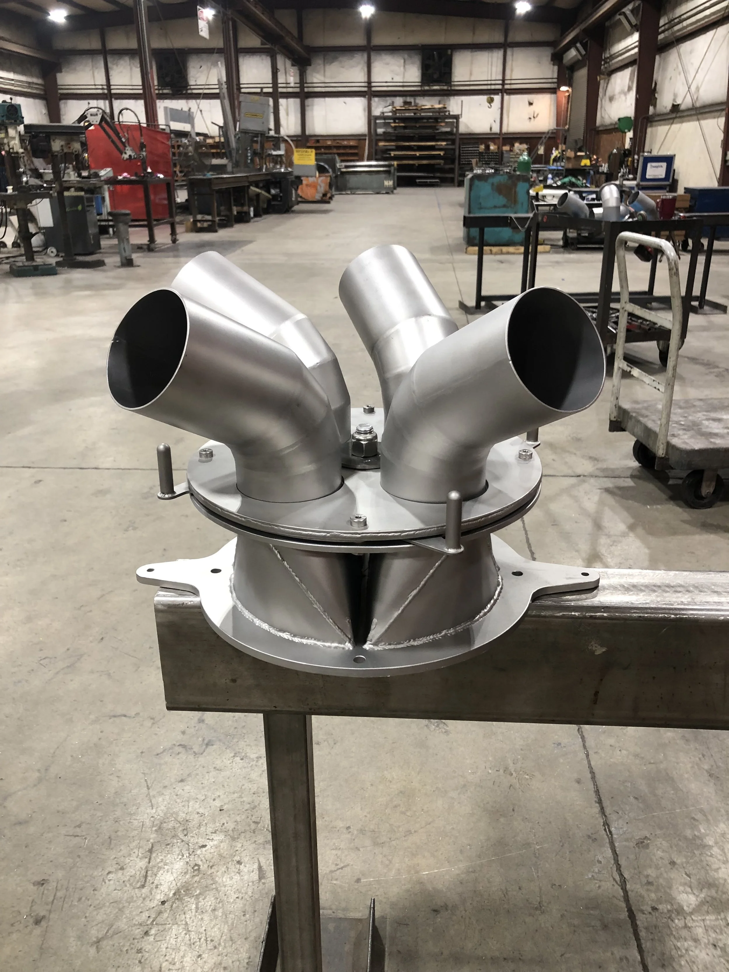 Metal exhaust or pipe components in a factory workshop with other machinery and workbenches in the background.