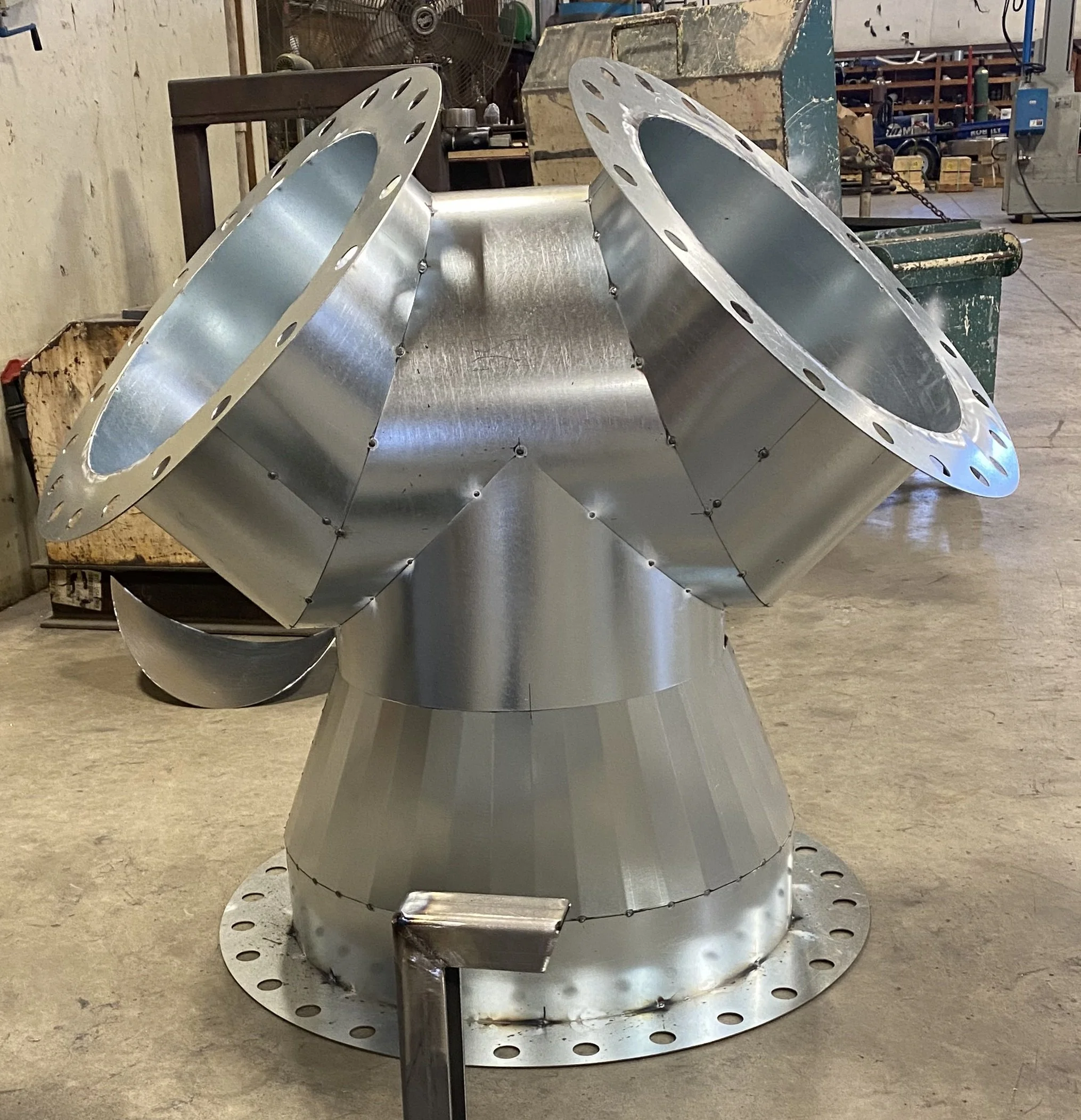 A large, metallic, industrial-looking structure made of welded metal sheets, resembling a machine or a piece of equipment, placed on a concrete floor in a workshop or factory setting.