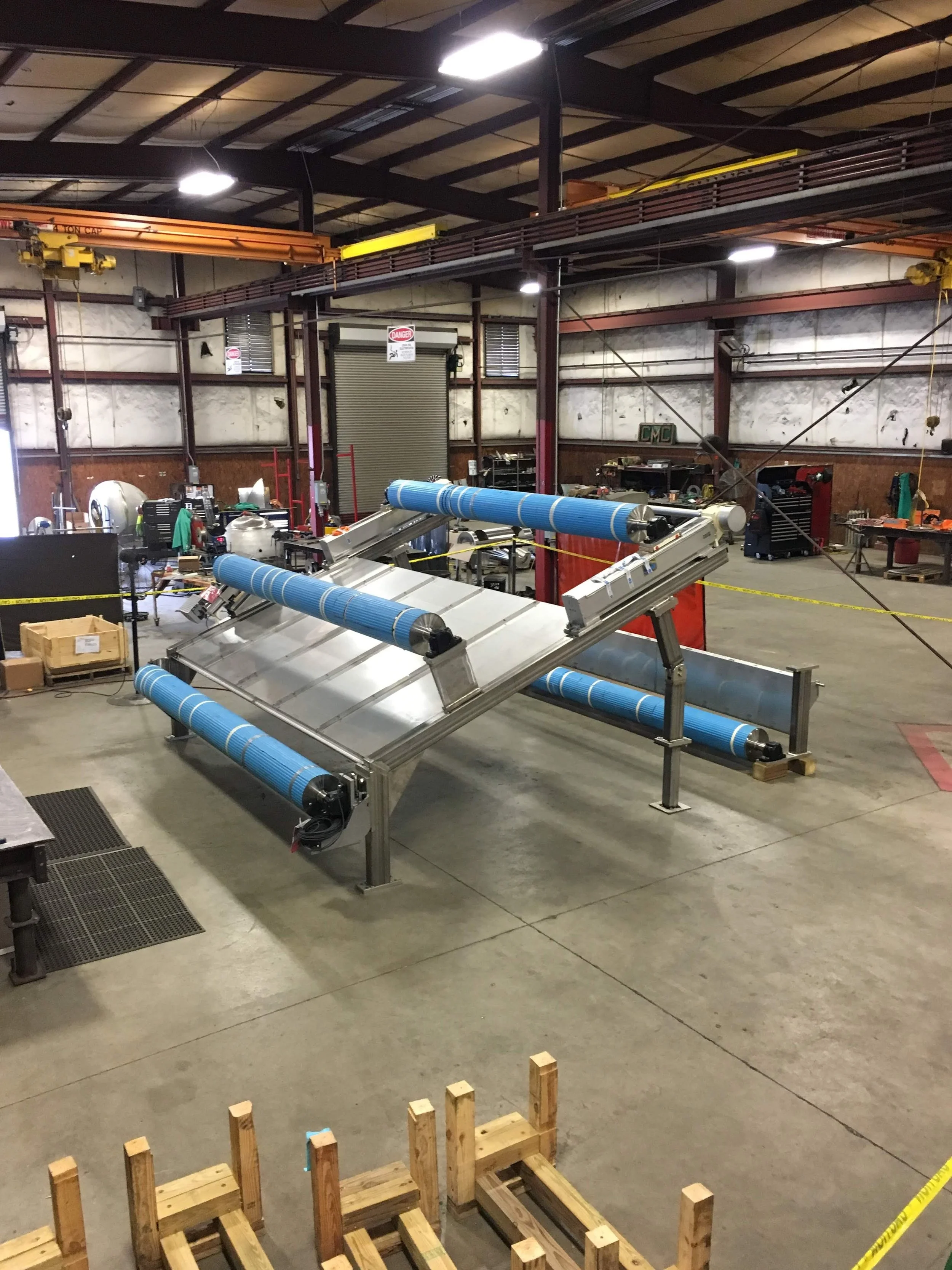 Industrial workshop with a large metal rolling machine with blue rollers, surrounded by various tools and equipment, and a staircase overhead.