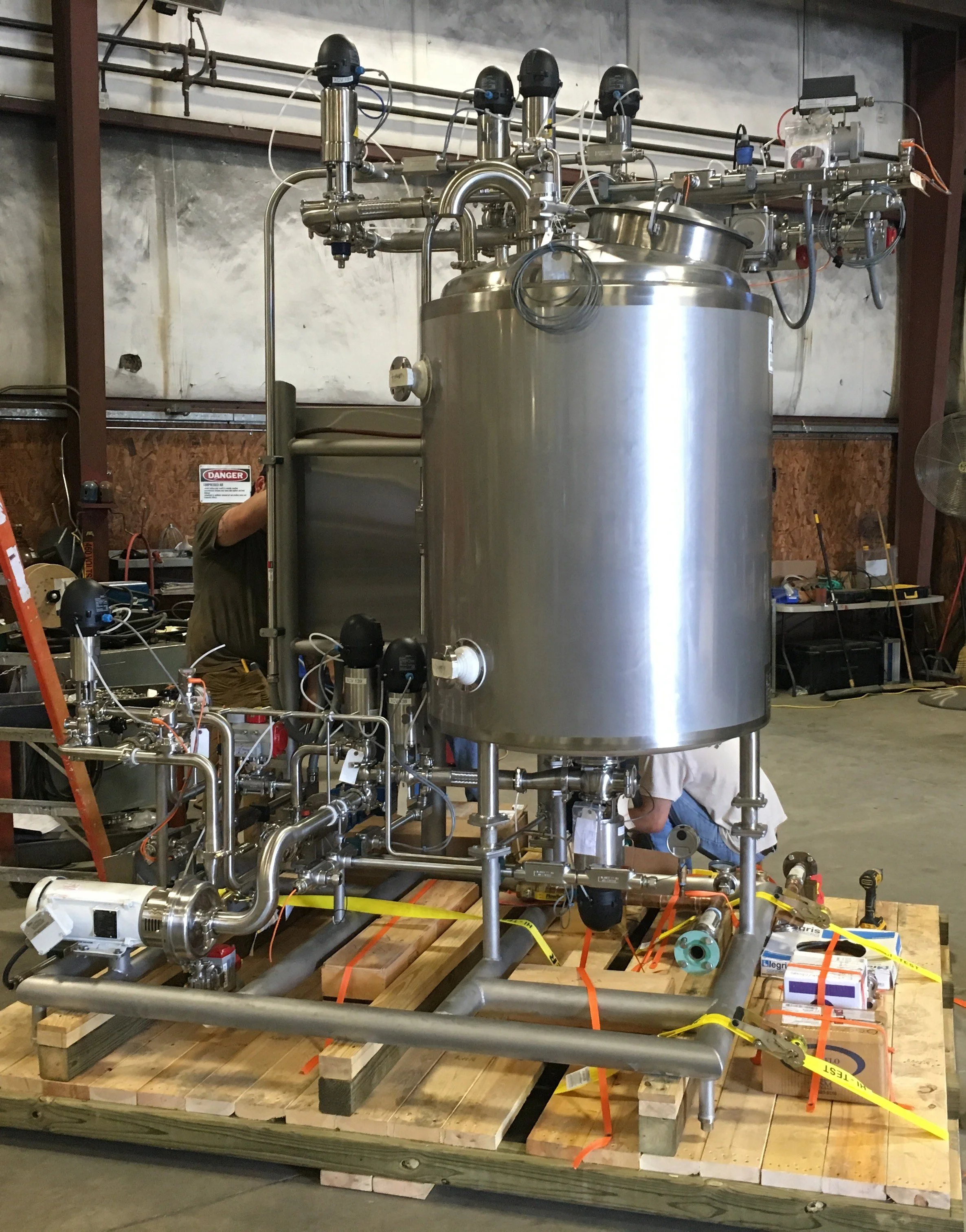 Large stainless steel industrial vessel with interconnected pipes, gauges, and controls, situated on a wooden pallet in a factory or workshop setting.