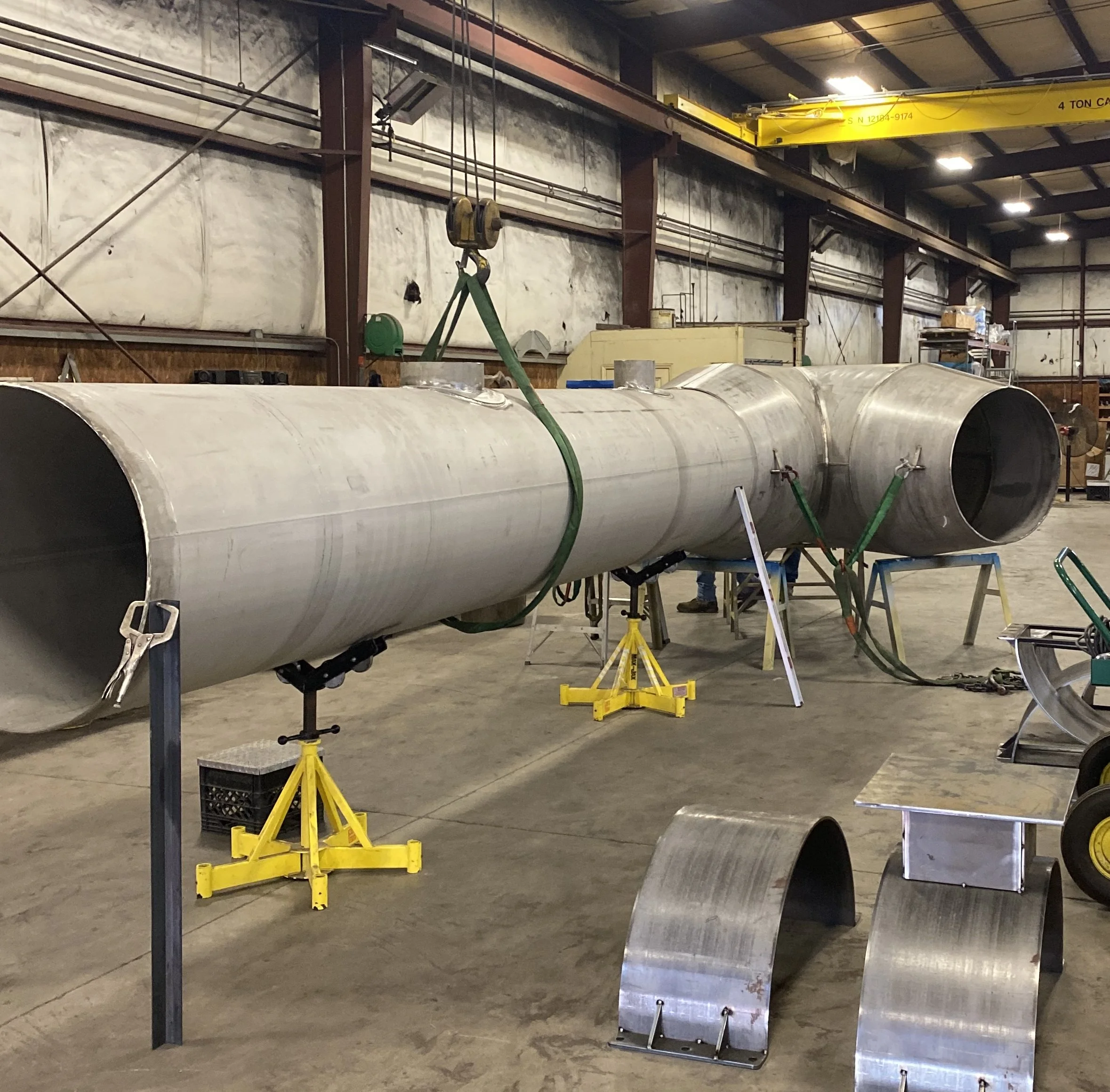 Large metal pipe in a workshop or industrial warehouse, supported by yellow stands, with equipment and tools nearby.