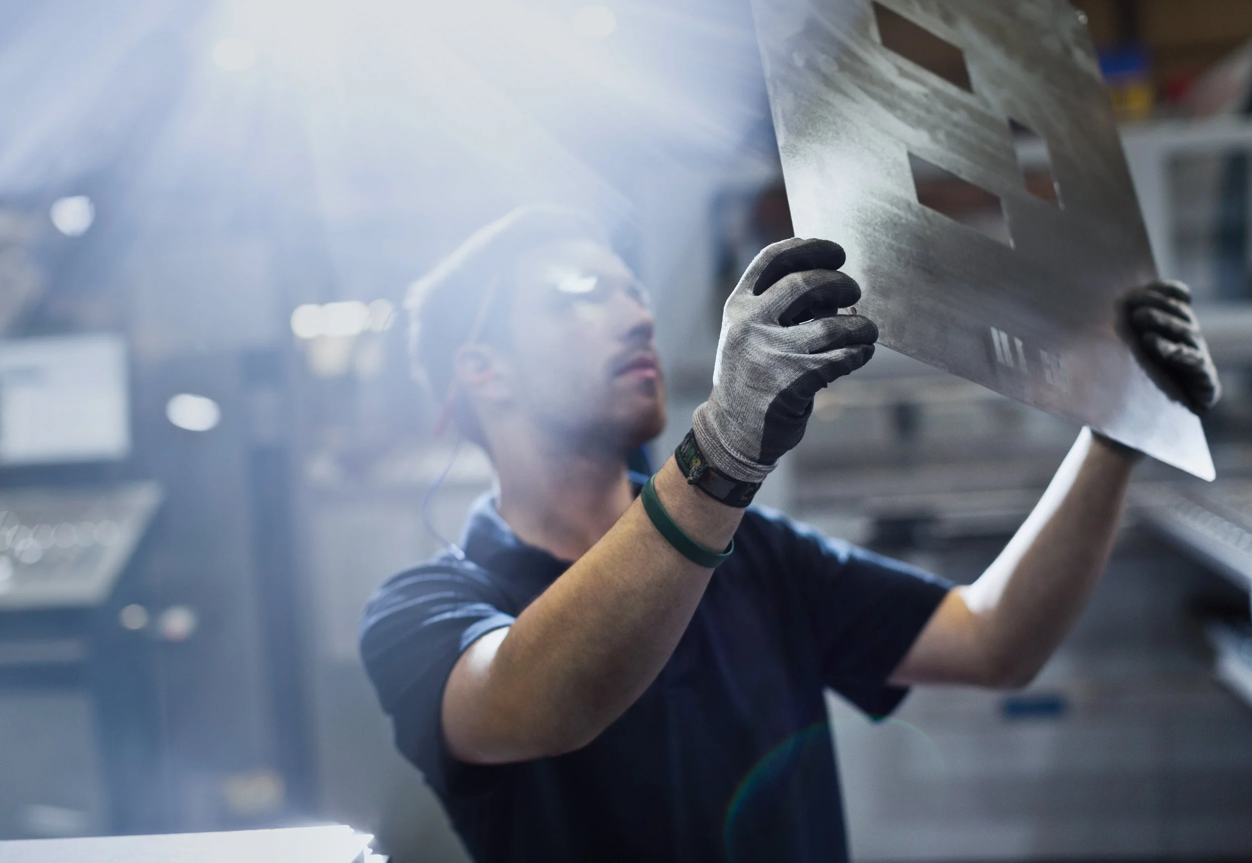A worker in a factory setting holding a metal sheet, wearing gloves and a cap, with sunlight shining from above.