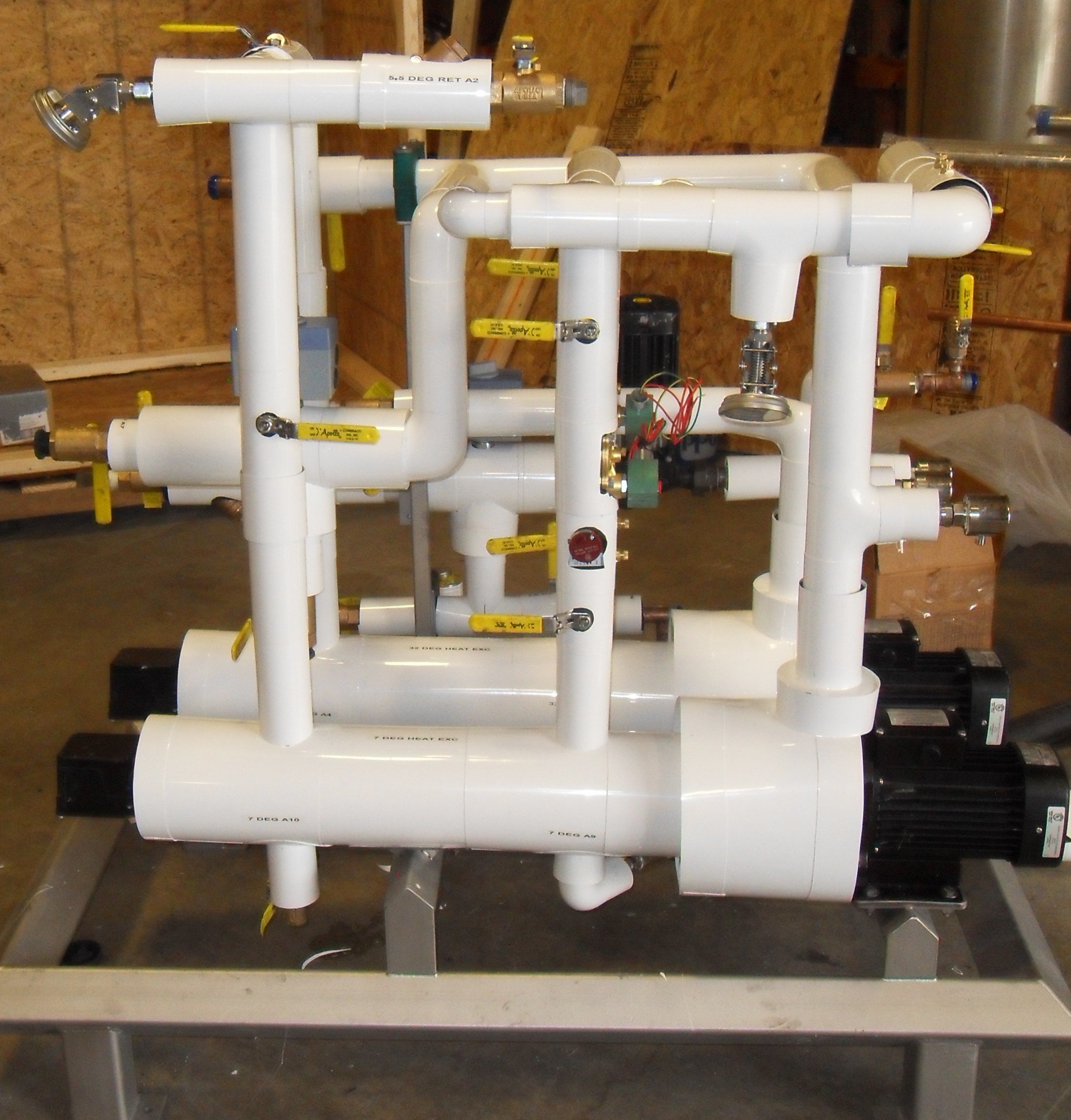 A complex system of white PVC pipes and valves assembled on a metal frame, with black motorized components at the base, installed indoors on a concrete floor.