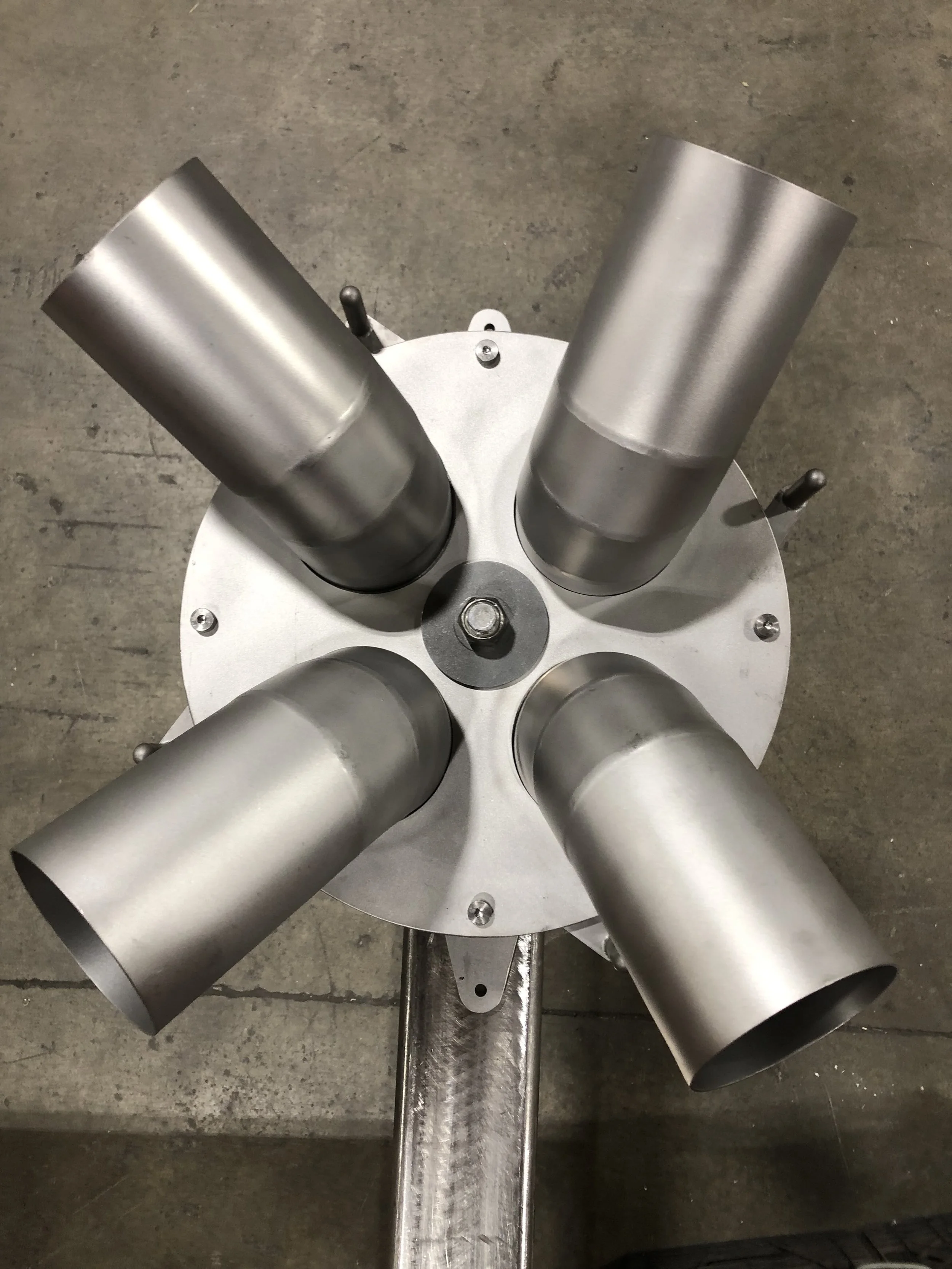 A metal fixture with four cylindrical openings arranged in a circular pattern on a round base, attached to a metal stand, on a concrete floor.