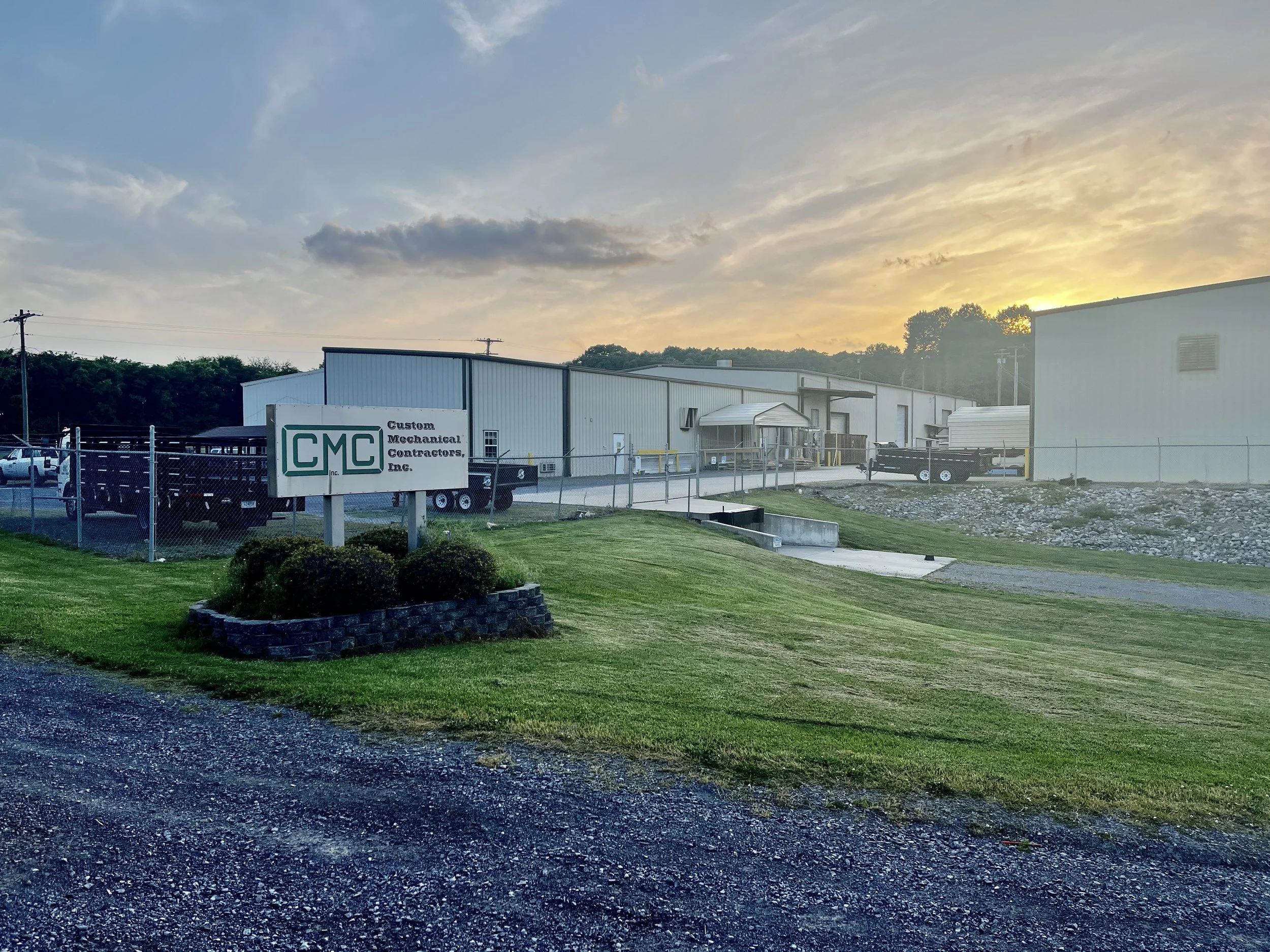 View of a commercial building complex with a sign that reads 'Custom Mechanical Contractors Inc.' at sunset, featuring a grassy area, gravel pathway, and parked trailers.