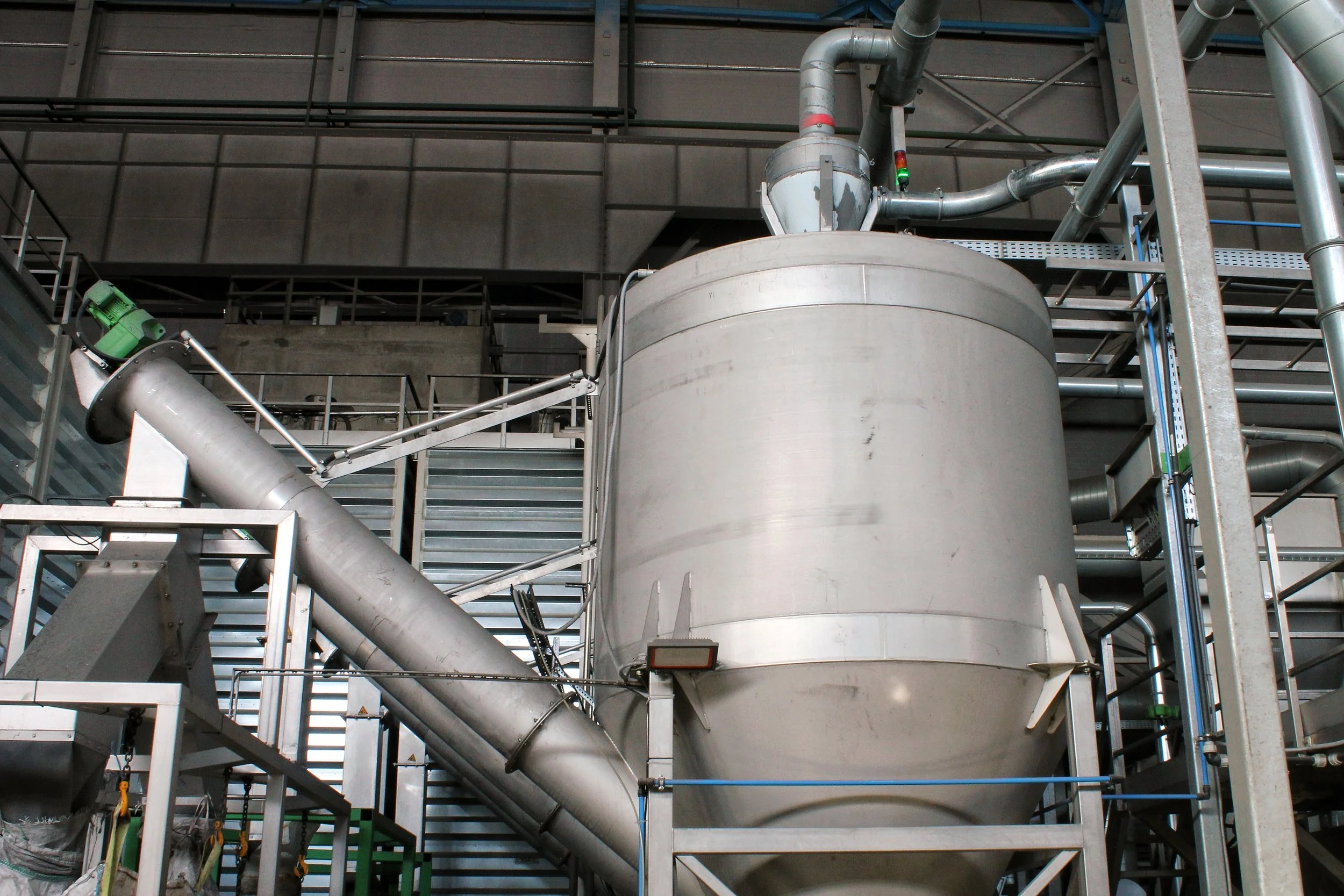Industrial equipment with large cylindrical metal tank, pipes, and conveyor in a factory setting.