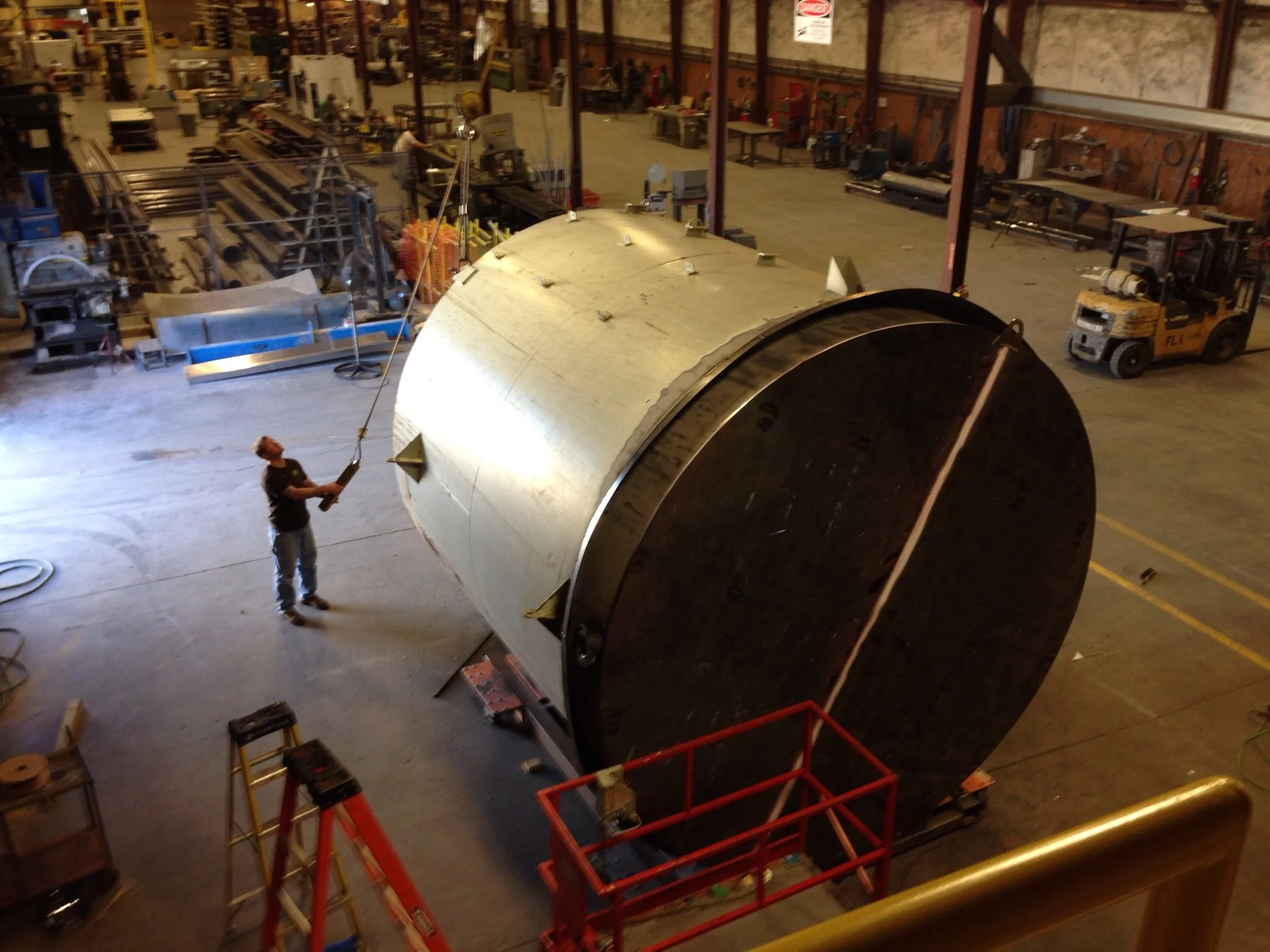 A person in a workshop is holding a tool and working on a large, cylindrical metallic object. The workshop is spacious with various equipment, tools, and machinery in the background.