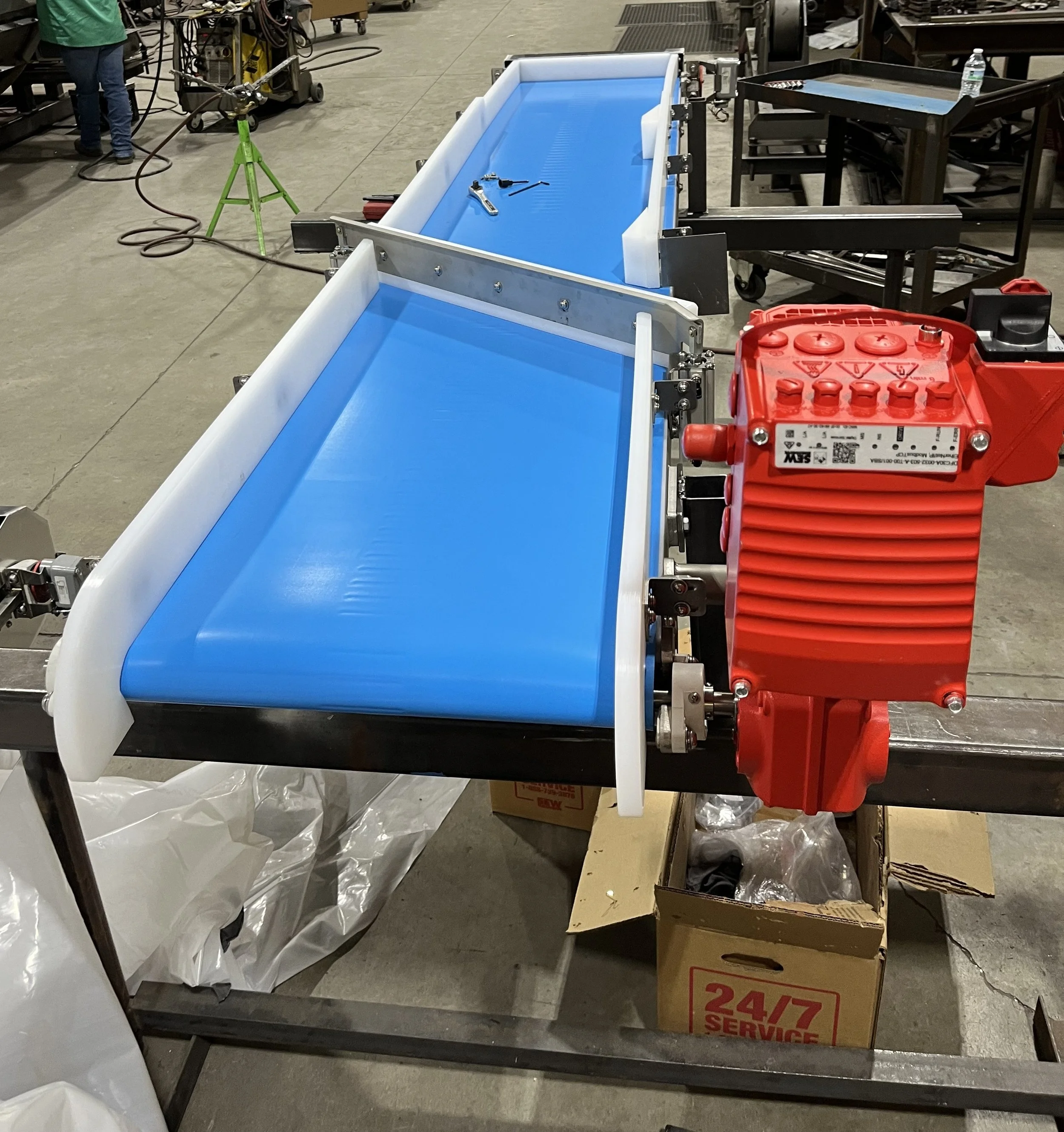 A conveyor belt with a blue surface and white side guards, equipped with a red motor at the end, in an industrial or manufacturing setting.