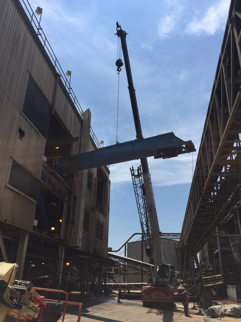 A crane lifting a large metal pipe between industrial warehouse buildings.