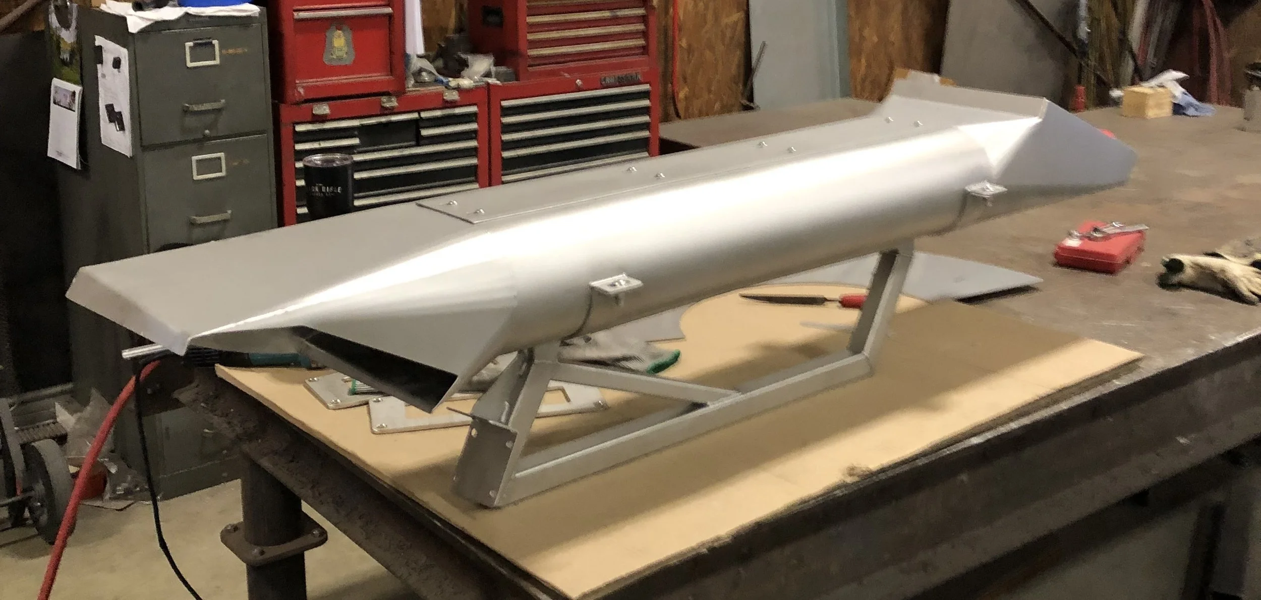 A metallic object on a workbench in a workshop, possibly an aircraft fuselage or a streamlined vehicle component, with tools and workshop equipment around.