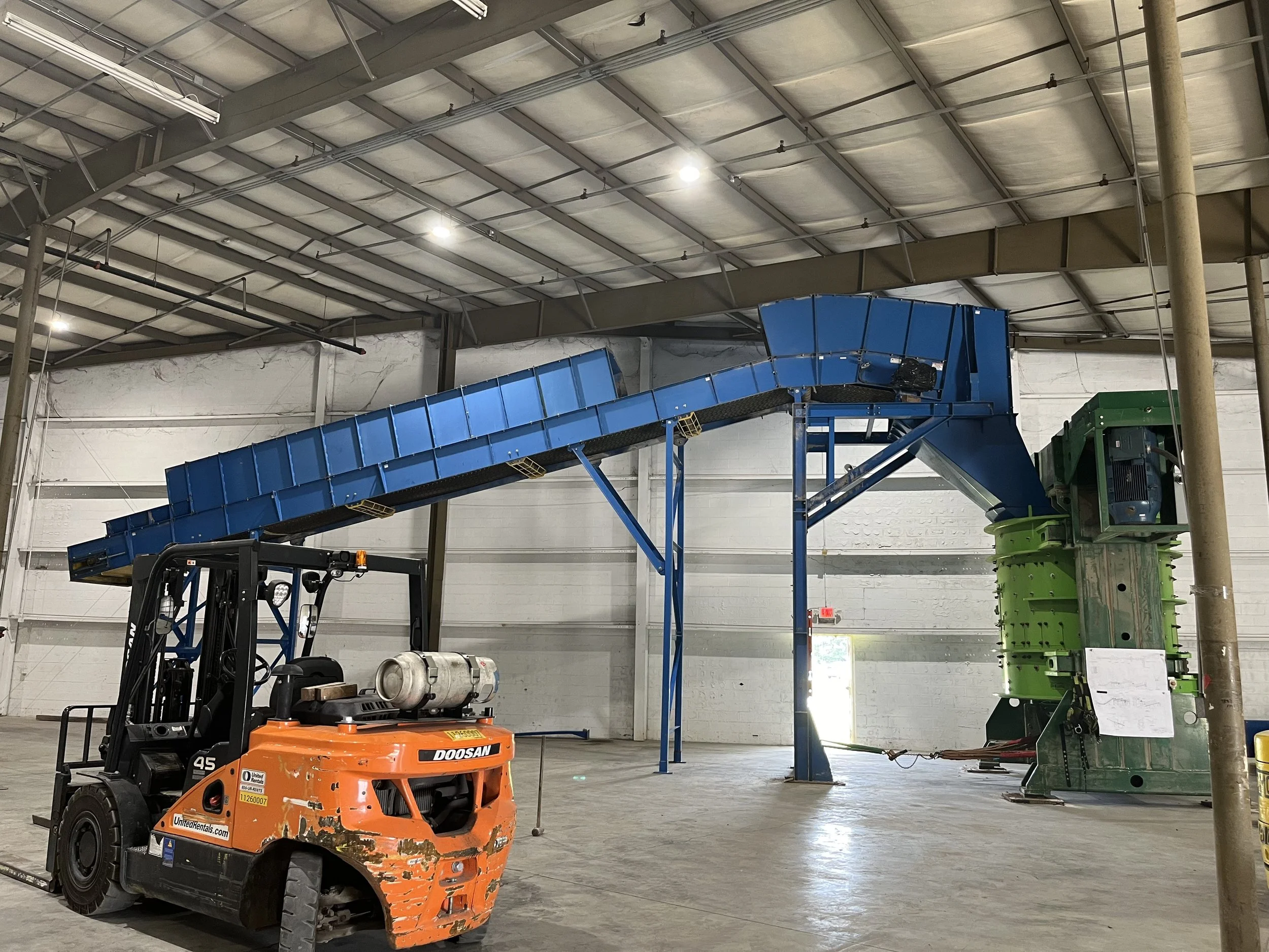 Industrial warehouse with blue conveyor system, green machinery, and orange forklift inside a spacious, well-lit building with metal ceiling and concrete floor.