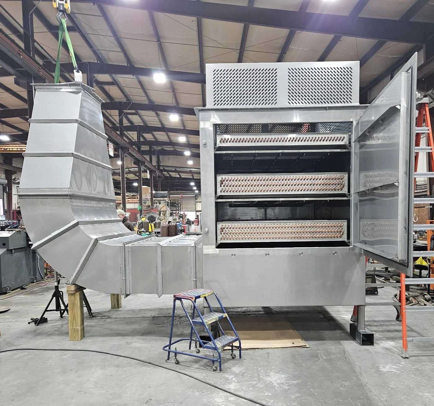 Large industrial stainless steel machine in a factory setting, with an open front view showing internal copper coils, connected to ductwork on the left, on a concrete floor with tools and equipment in the background.