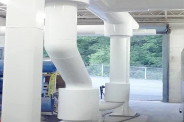 Indoor space with large white industrial-looking ducts and pipes, and an open garage-style door revealing greenery outside.