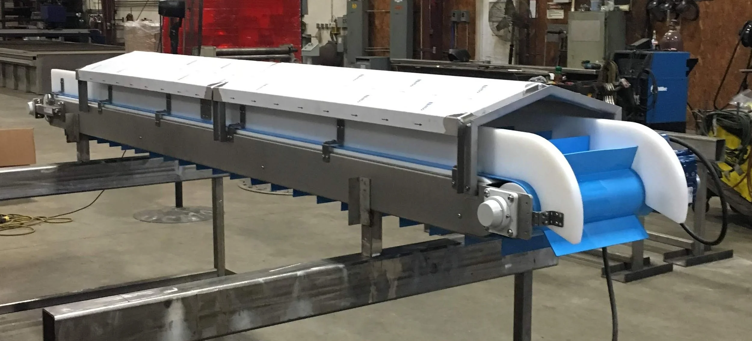 Industrial conveyor system in a workshop setting with metal components and machinery in the background.