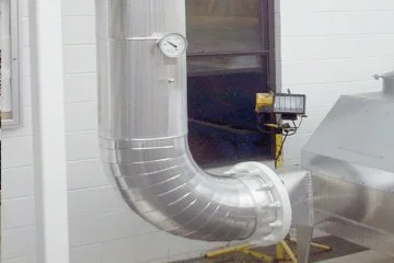 Metal duct pipe with a gauge mounted on it, in an industrial setting with a white painted brick wall.