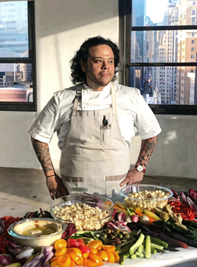 Chef with curly hair and tattoos wearing a white apron, standing beside a table filled with bowls of snacks and vegetables in a bright, urban studio with large windows.