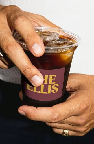 Person holding a plastic cup of dark soda with ice, labeled 'The Ellis'.