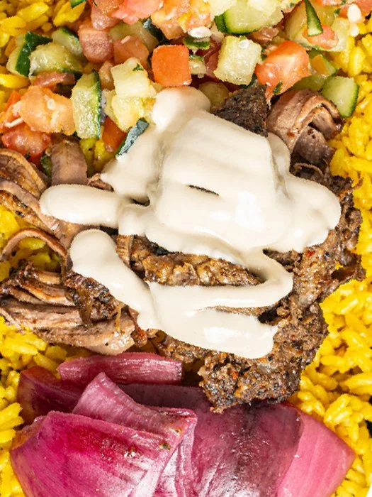 Close-up of a plate of Mexican food featuring grilled meat topped with white sauce, surrounded by yellow rice, chopped cucumber, tomato, onion, and diced avocado, with some pickled red onions at the bottom.