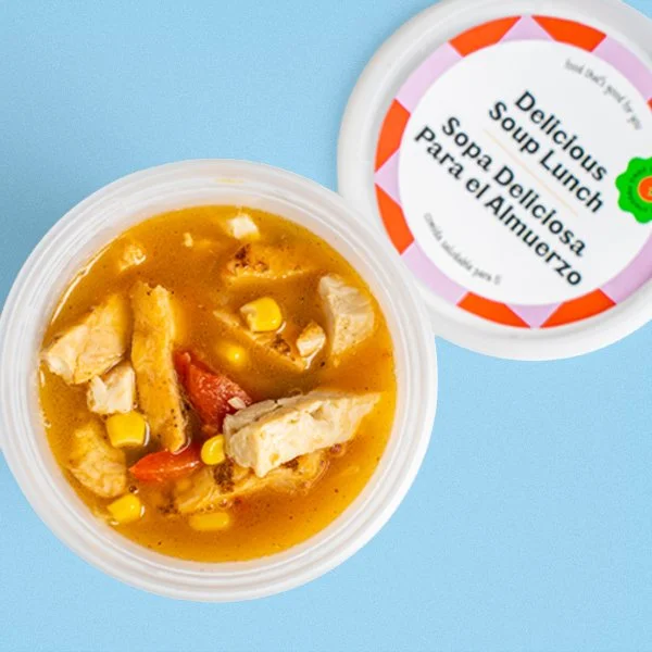 Container of chicken soup with vegetables and broth, next to a lid labeled 'Delicious Soup Lunch' in multiple languages.