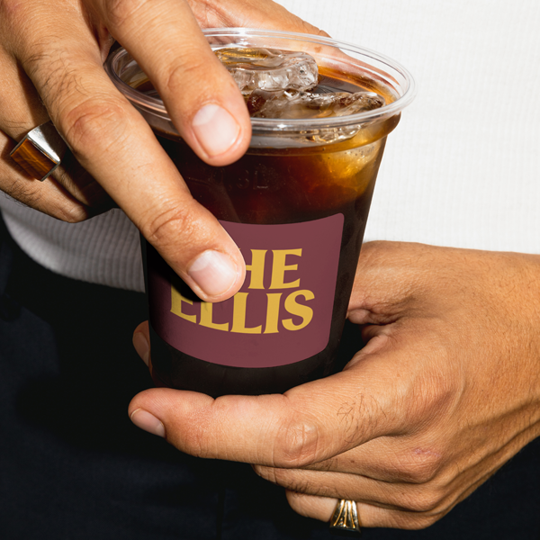 Person holding a plastic cup of iced coffee with a custom label that reads "The Ellis."