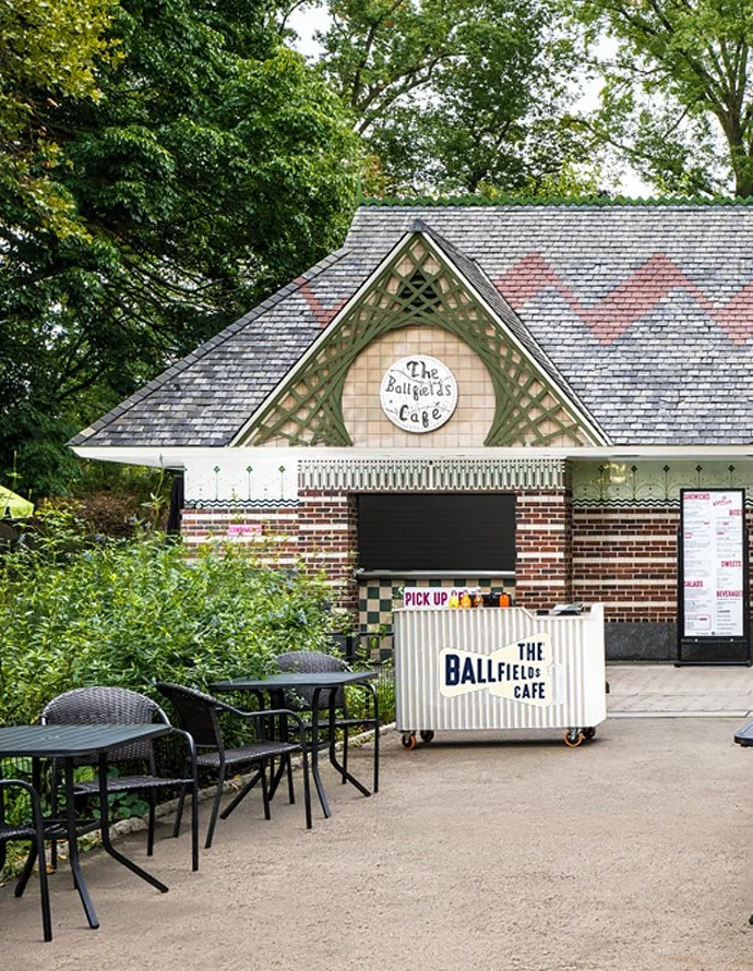 Exterior of The Ballfields Cafe with outdoor seating, a pick-up cart, and lush green trees in the background.