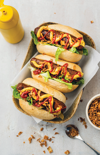 Three hot dogs topped with mustard, ketchup, and sautéed onions and peppers in a basket lined with white paper, with a yellow mustard bottle and a small bowl of crispy fried onions nearby.