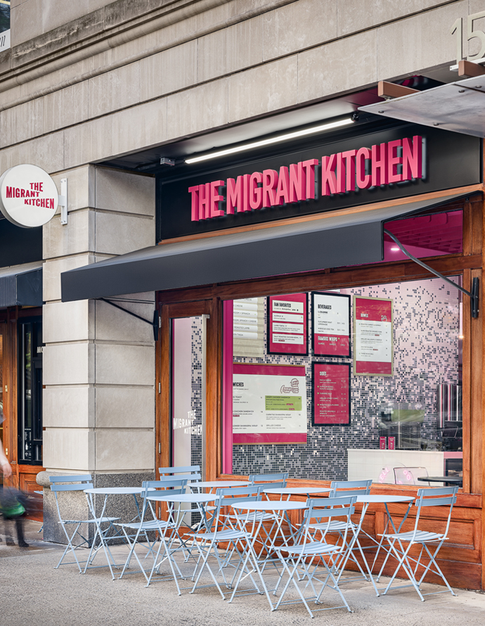 Exterior of a restaurant called 'The Migrant Kitchen' with pink illuminated sign, outdoor seating with white tables and chairs, and menu displayed in the window.