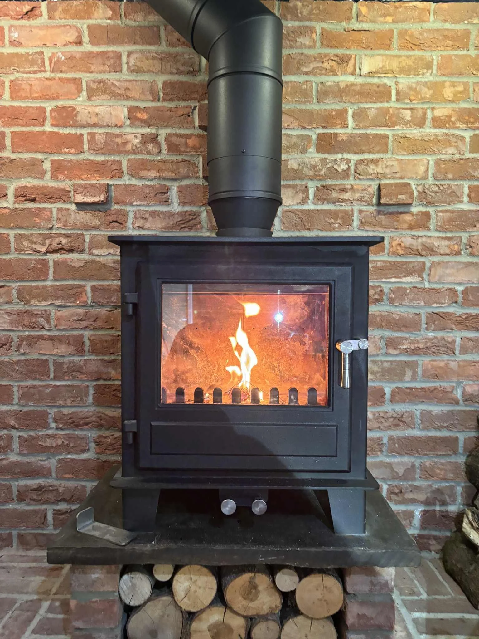 A black wood-burning stove with a glass door showing a fire inside, surrounded by a brick wall, with logs stored underneath.