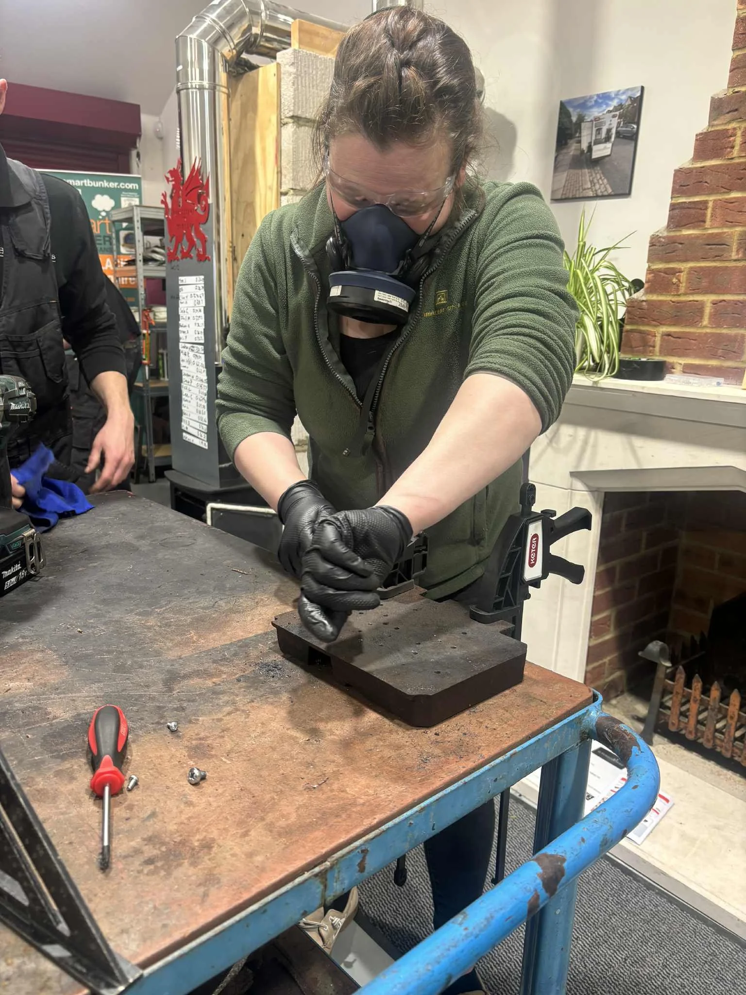 Woman wearing a green jacket, gloves, and a mask working with a clamp on a piece of black material at a workshop table.