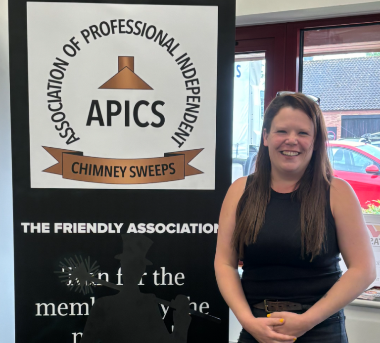 A woman standing next to a sign that reads 'APICS, The Friendly Association, Chimney Sweeps' with a logo of a chimney.