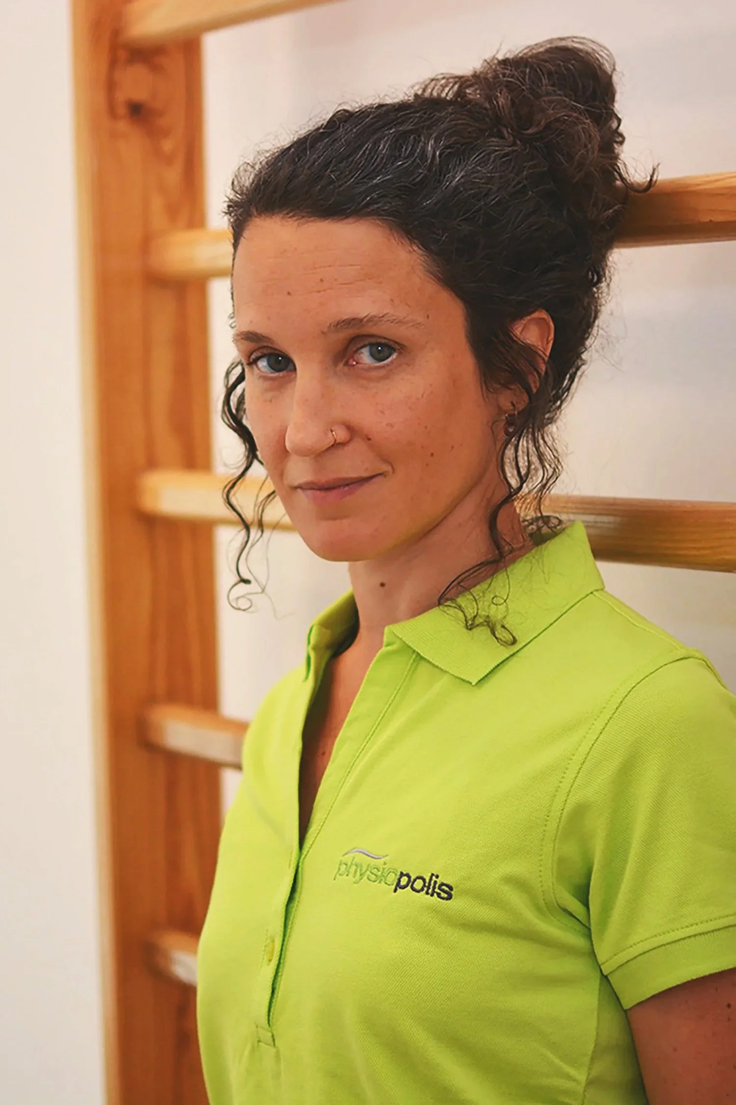 Junge Frau mit kurzen dunklen Haaren und Nasenpiercing trägt ein leuchtend grünes Hemd mit Aufdruck 'physiopolis' und steht vor einem Holz-Gitter im Innenbereich.
