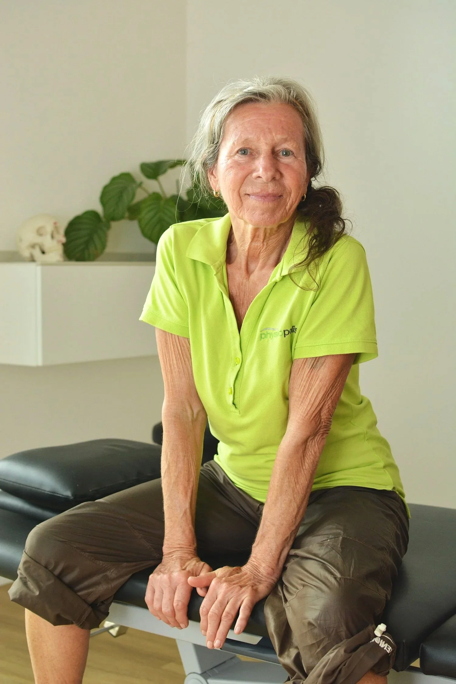 Ältere Frau in einem grünen Polo sitzt auf einer Behandlungsliege in einer Praxis für Physiotherapie.