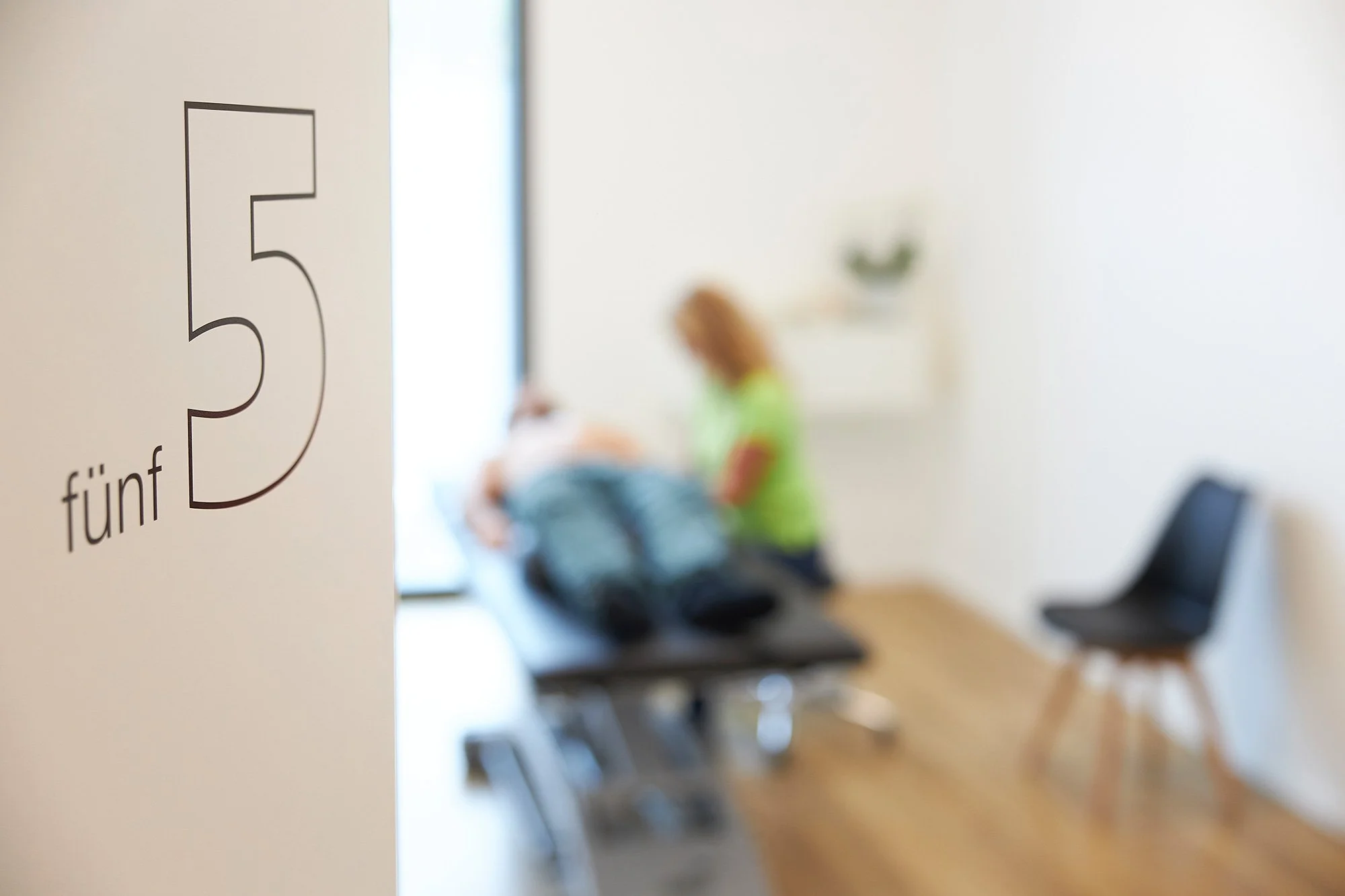 Ein gekachelter Raum mit einem weißen Wandanstrich, auf dem ein Vorhängeschloss an einem Türgriff angebracht ist. Im Hintergrund sieht man eine Behandlungsliege, auf der eine Person liegt, sowie eine Frau in grünem Hemd, die mit ihr spricht. Es gibt 