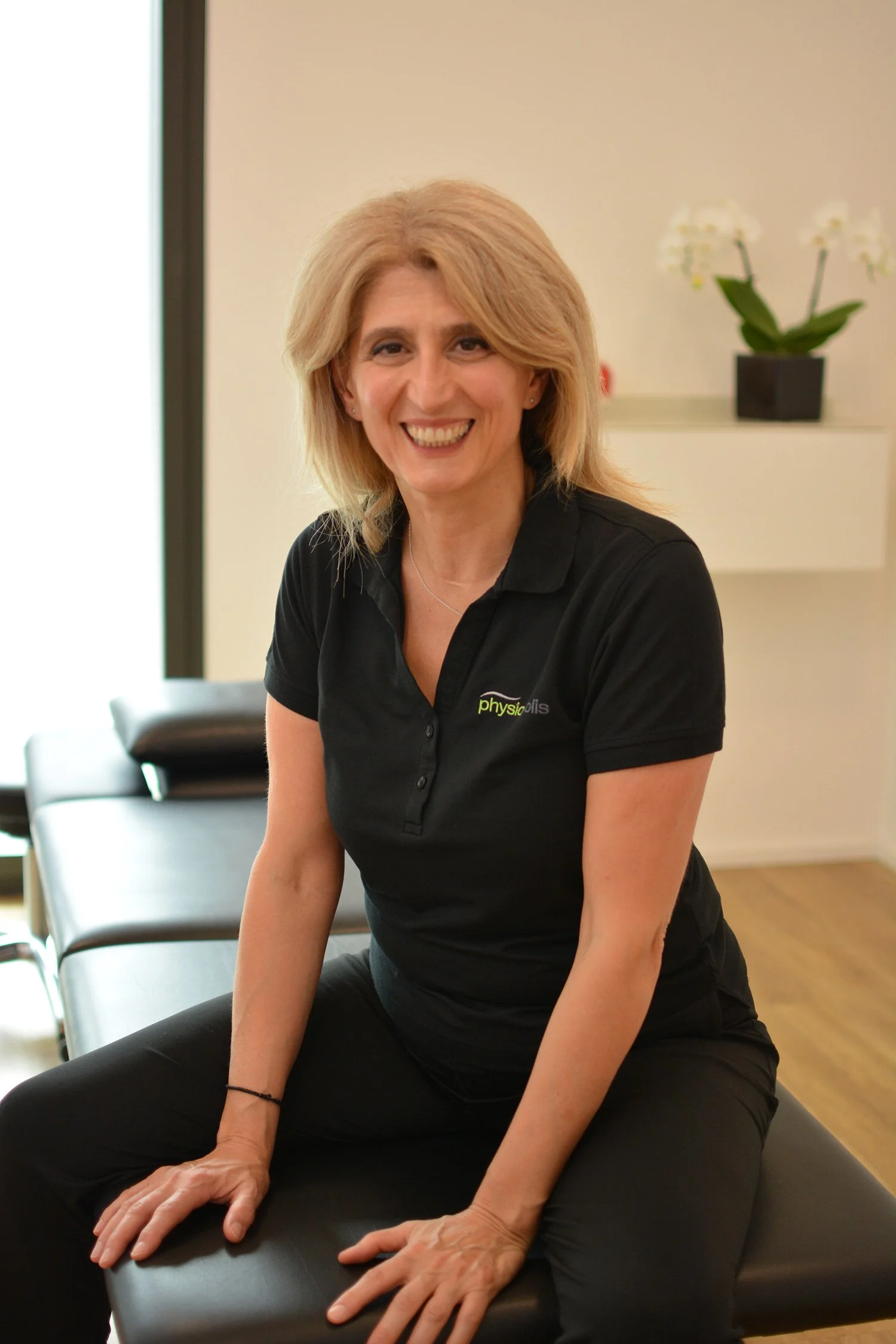 Eine lachende Frau sitzt auf einer schwarzen Behandlungsliege in einem Raum mit weißer Wand und einer weißen Kommode mit einer Topfpflanze.