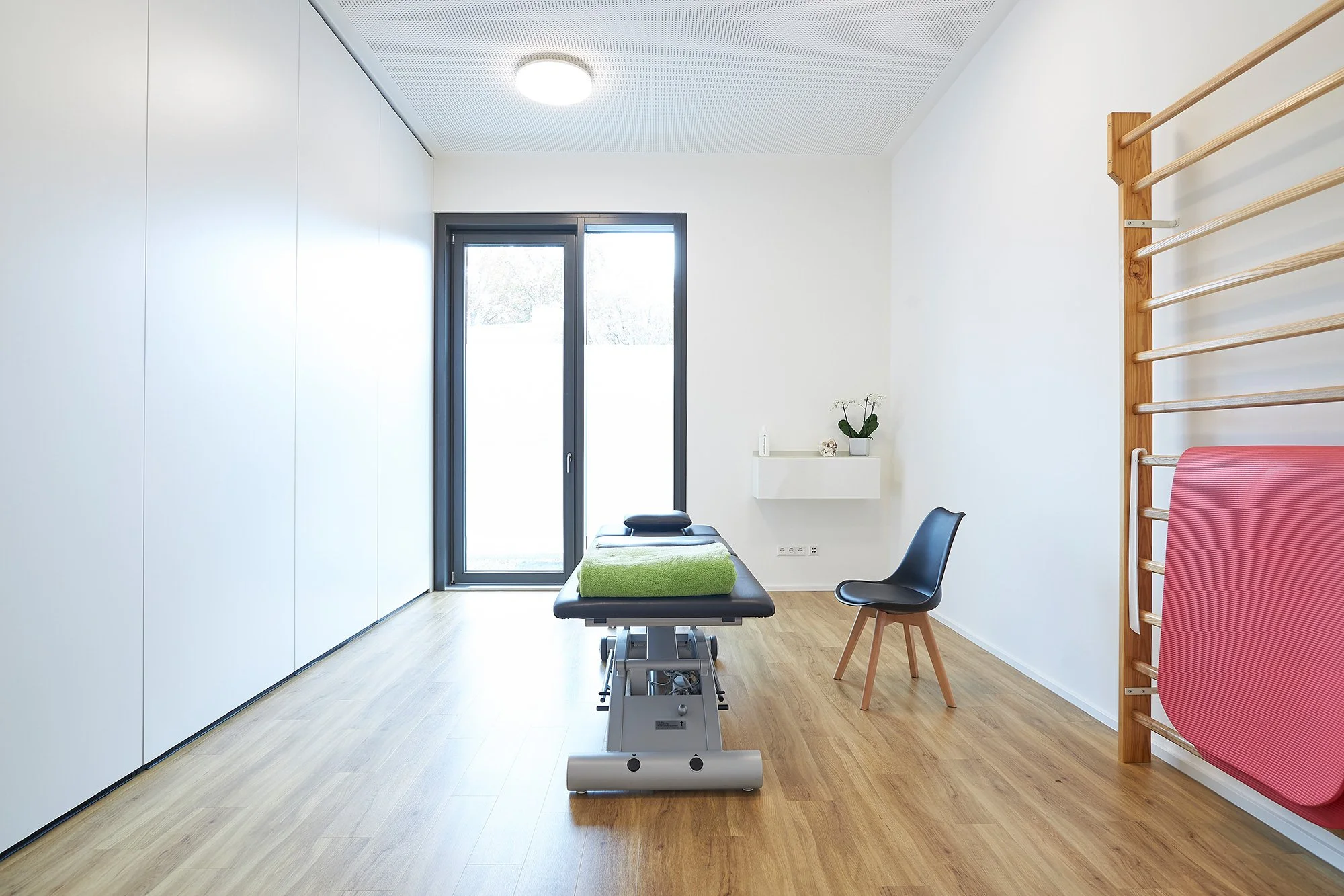 Behandlungsraum mit Behandlungsliege, Stuhl, Wandregal, Gipswand, Fenster und Holzwand mit Kletterstange