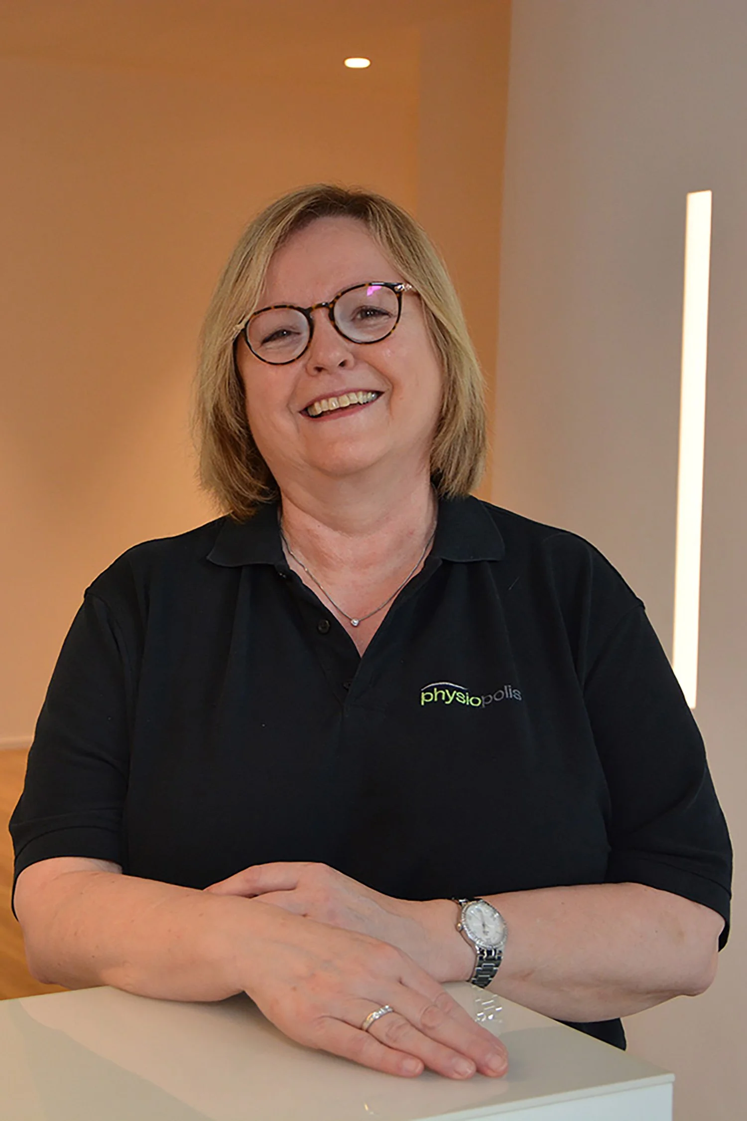 Eine lächelnde Frau mit Brille, schwarzem Polo-Shirt mit 'physiopolis'-Logo, sitzt an einem weißen Tisch.