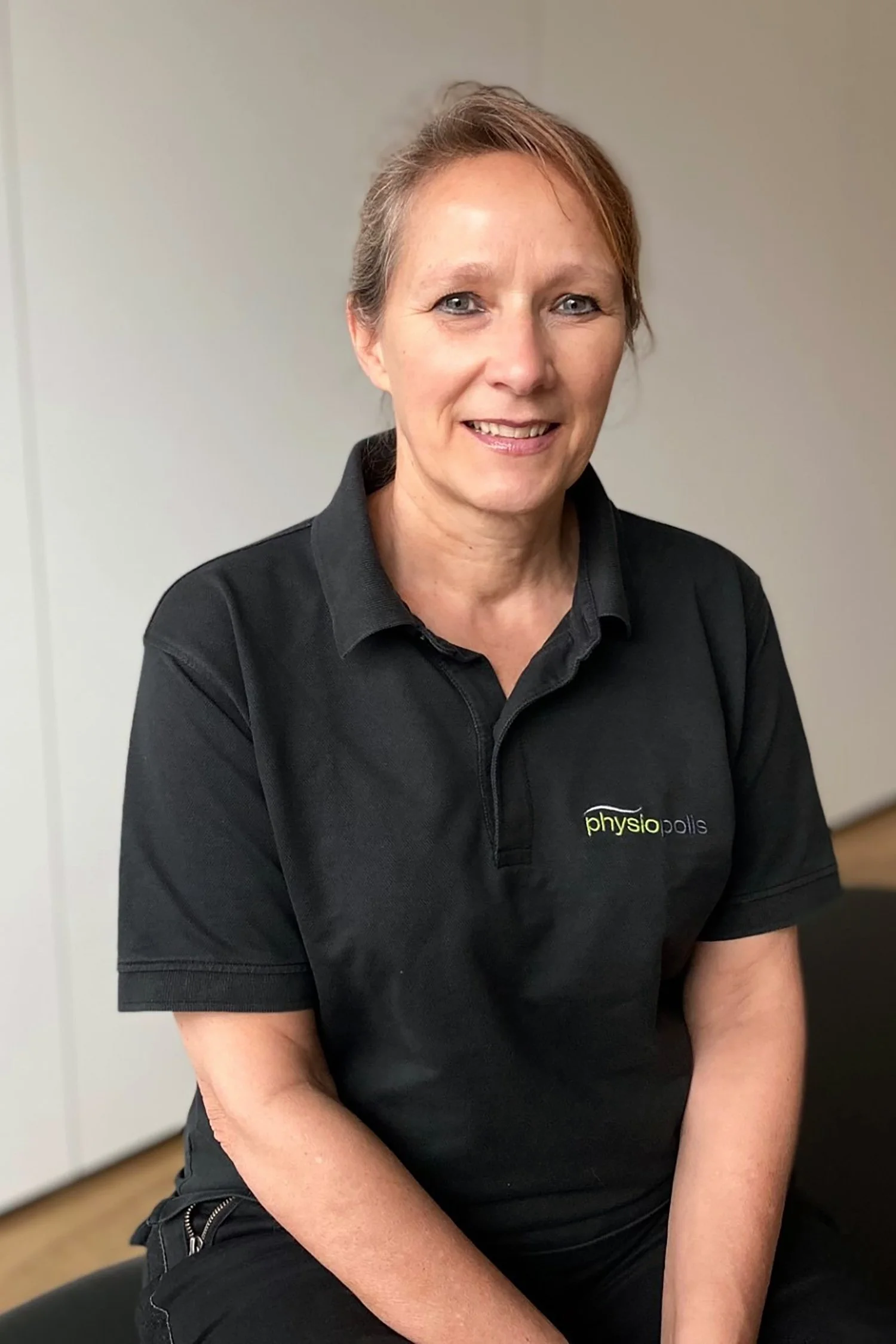 Ein Porträt einer lächelnden Frau mit kurzen, braunen Haaren in einem schwarzen Polohemd mit dem Logo "physio polis" auf der Brust, sitzend in einem Raum mit neutralem Hintergrund.