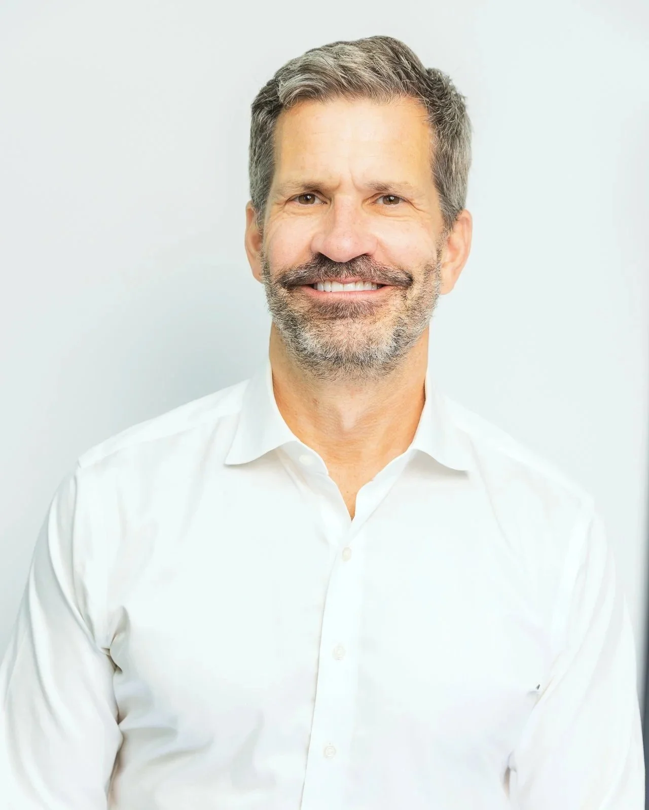 A smiling middle-aged man with a beard and gray hair wearing a white dress shirt against a plain background.