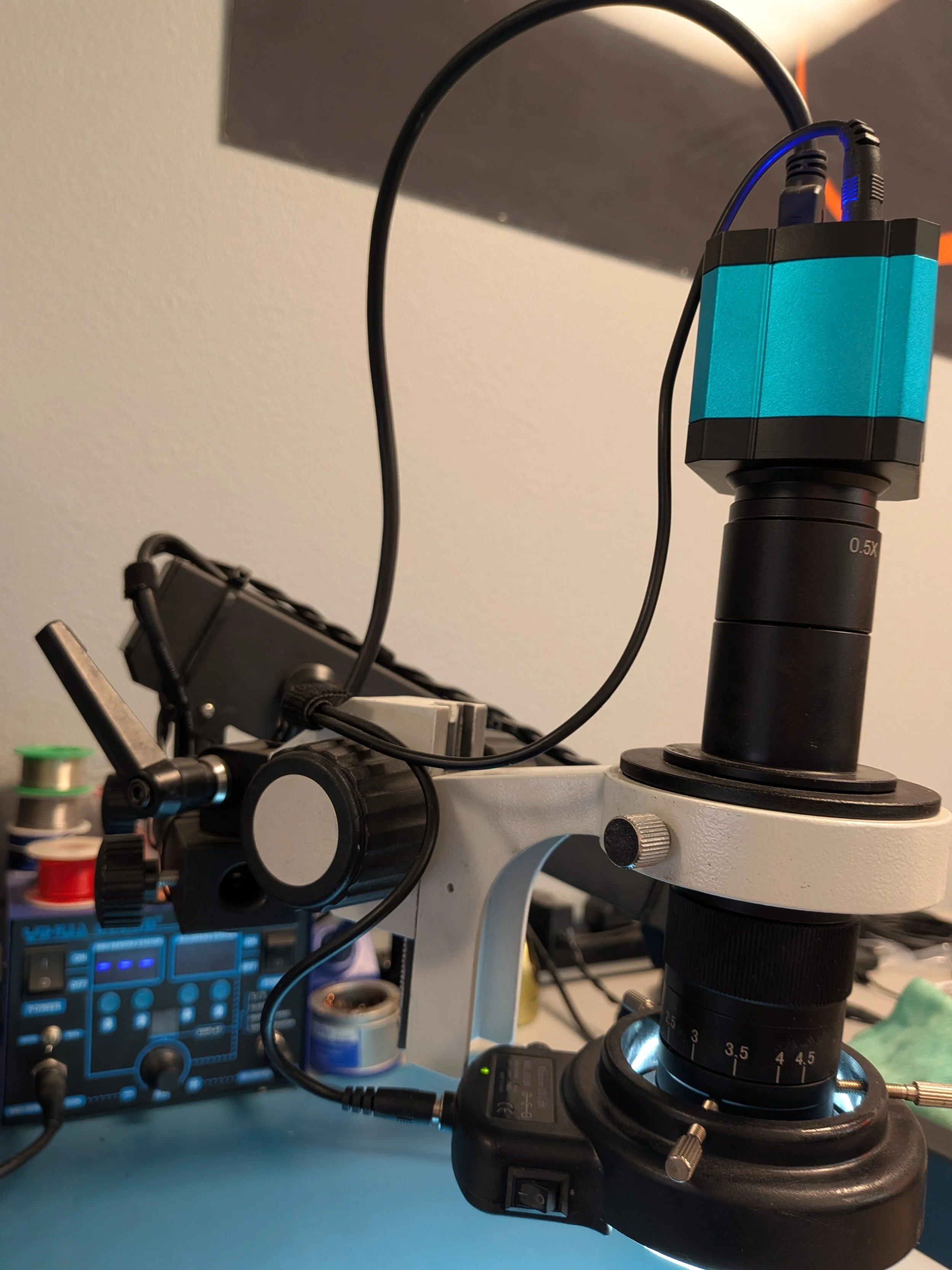 Close-up of a white microscope with a black eyepiece and a blue and black camera mounted on top. In the background, there is a blue electronic device and some spools of wire.