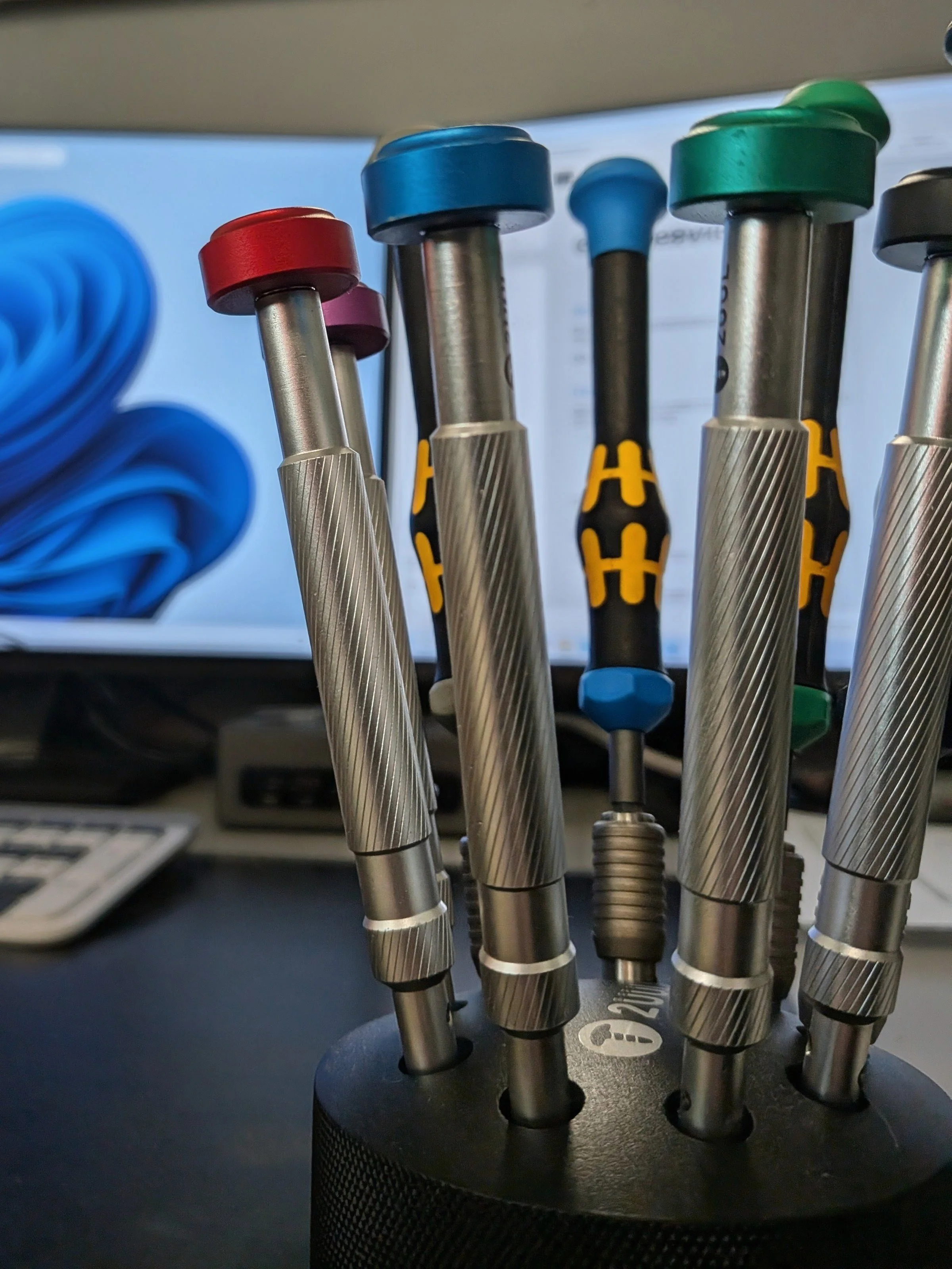 Set of precision screwdrivers with colored handles on a black rotating stand, with a computer monitor in the background.
