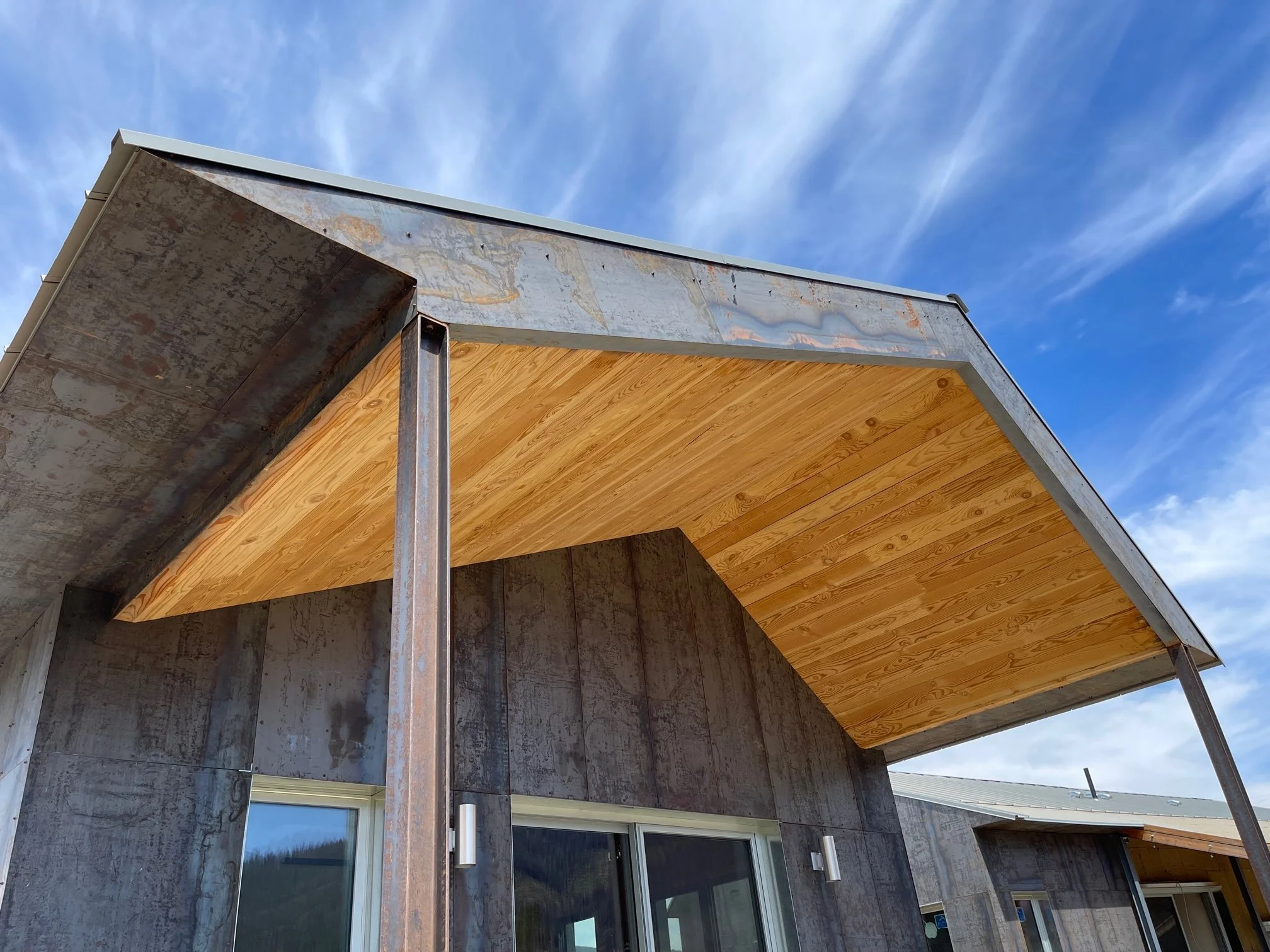 Mass Timber home exterior with a steel panel exterior, wooden underside exposed on the porch roof, and blue sky with clouds.