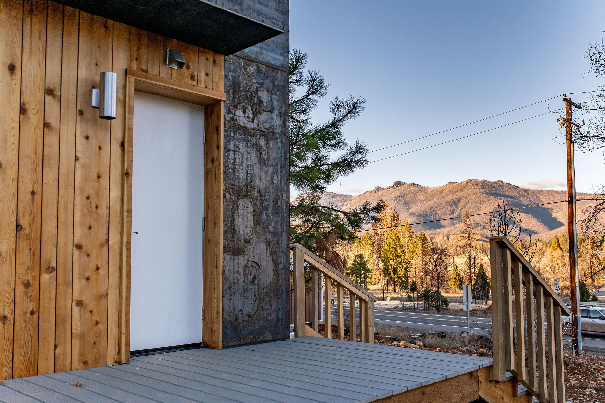 Close-up of a modern CLT building's exterior, featuring a white door with wooden trim, surrounded by CLT paneling and a steel wall, with a wooden staircase leading up to it, set against a scenic mountainous landscape and a clear blue sky.