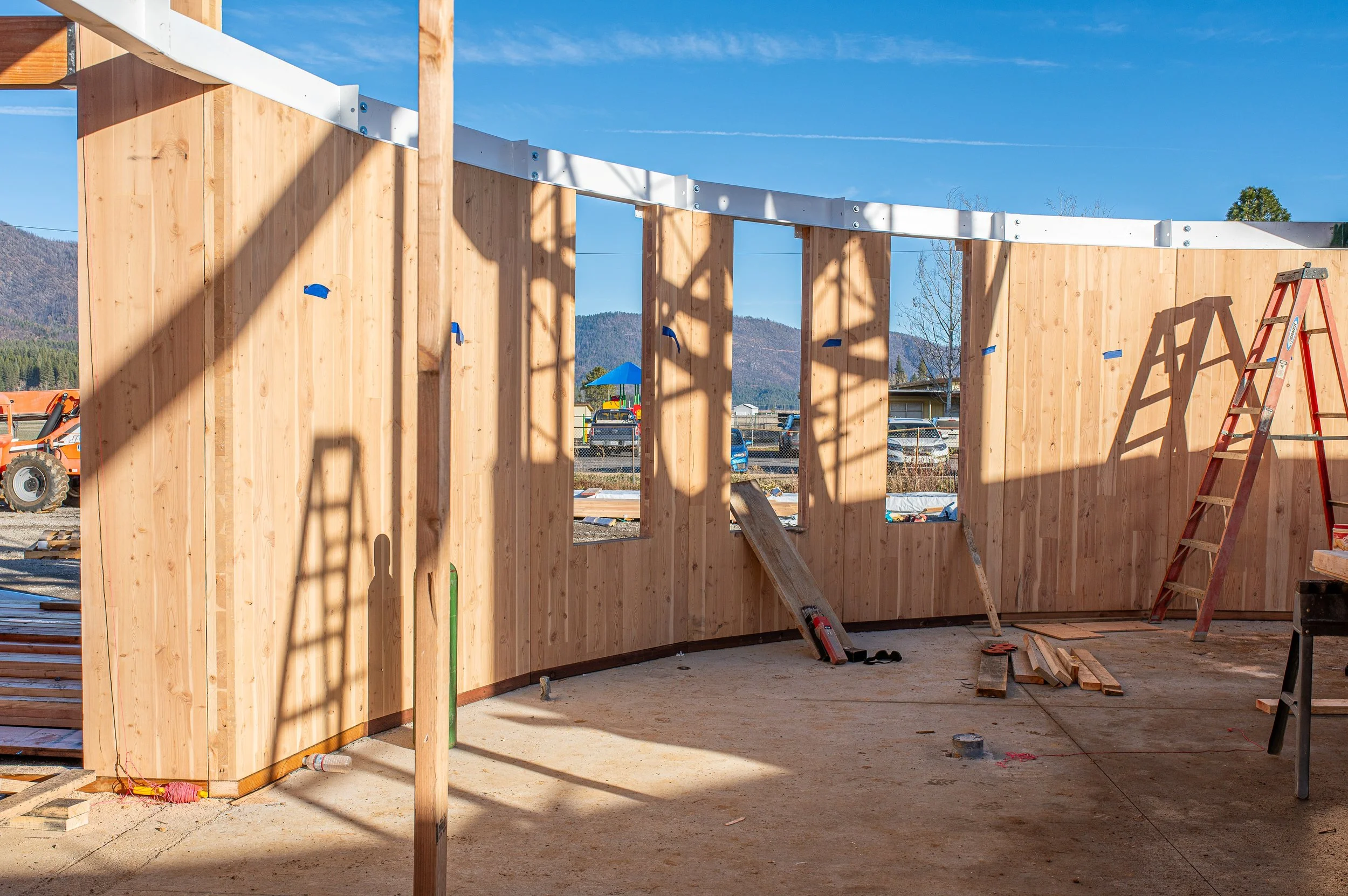 Maidu Roundhouse Council building, CLT wooden wall framing under construction with ladders, construction tools, and equipment on site. Clear blue sky and distant mountains in background.