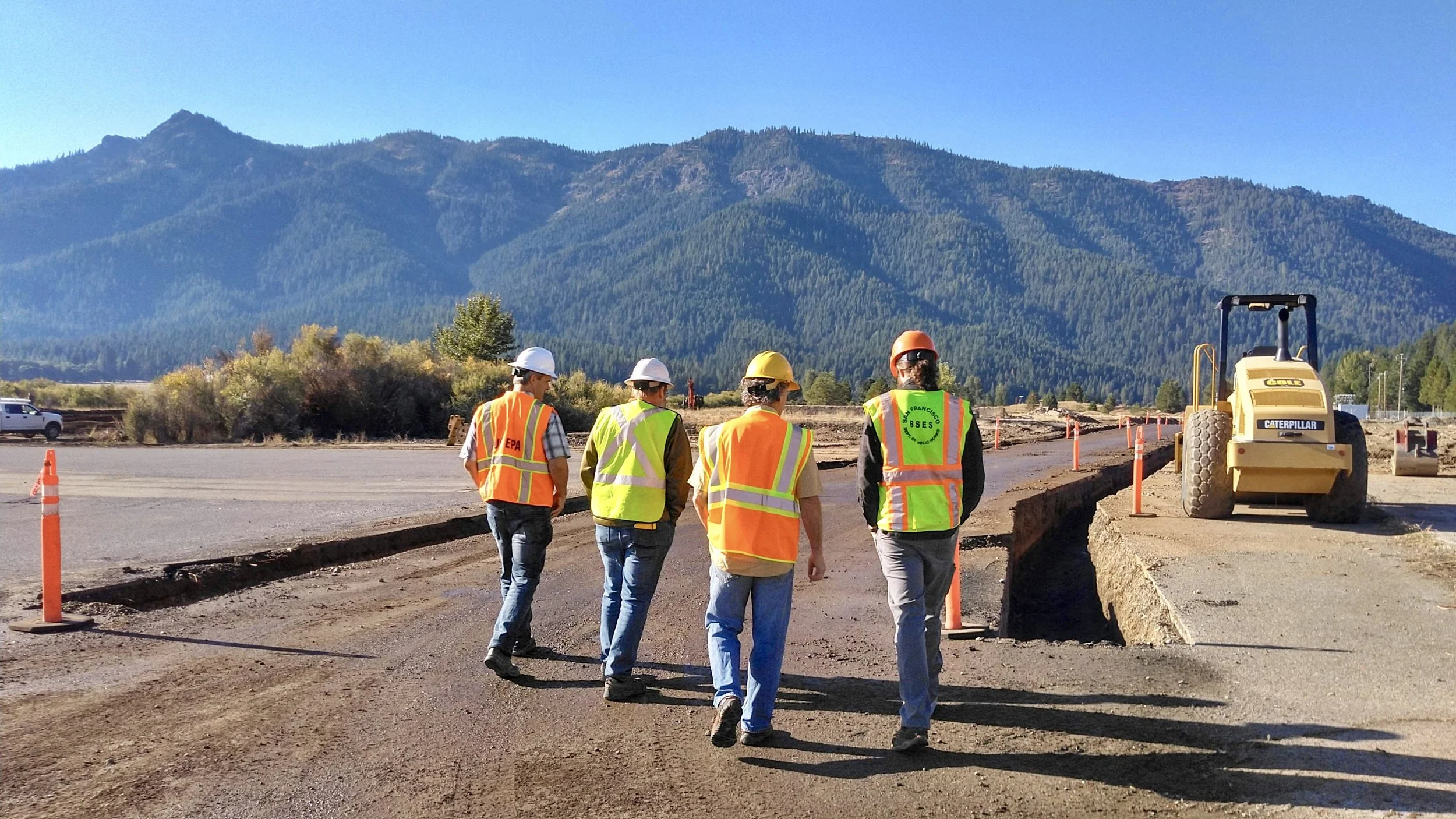 Construction workers wearing safety vests and helmets walking on a construction site with mountains in the background.