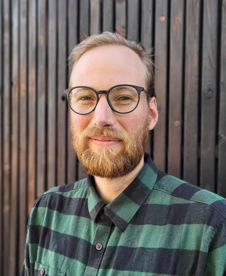 Nicholas Sills, A man with glasses, a beard, and light brown hair posing outdoors in front of a dark wooden fence, wearing a green and black plaid shirt.