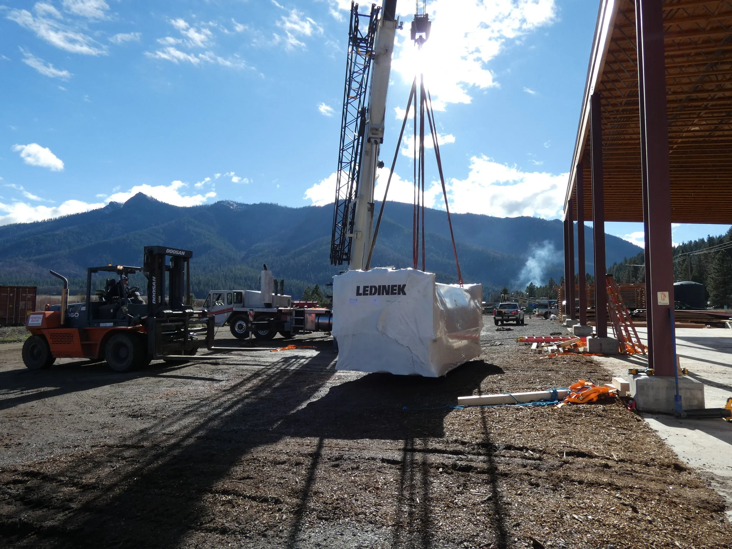 Crescent Mills, CA facility site with a crane lifting a large, wrapped piece of fabrication equipment, a forklift, and construction vehicles, with a mountain landscape in the background.