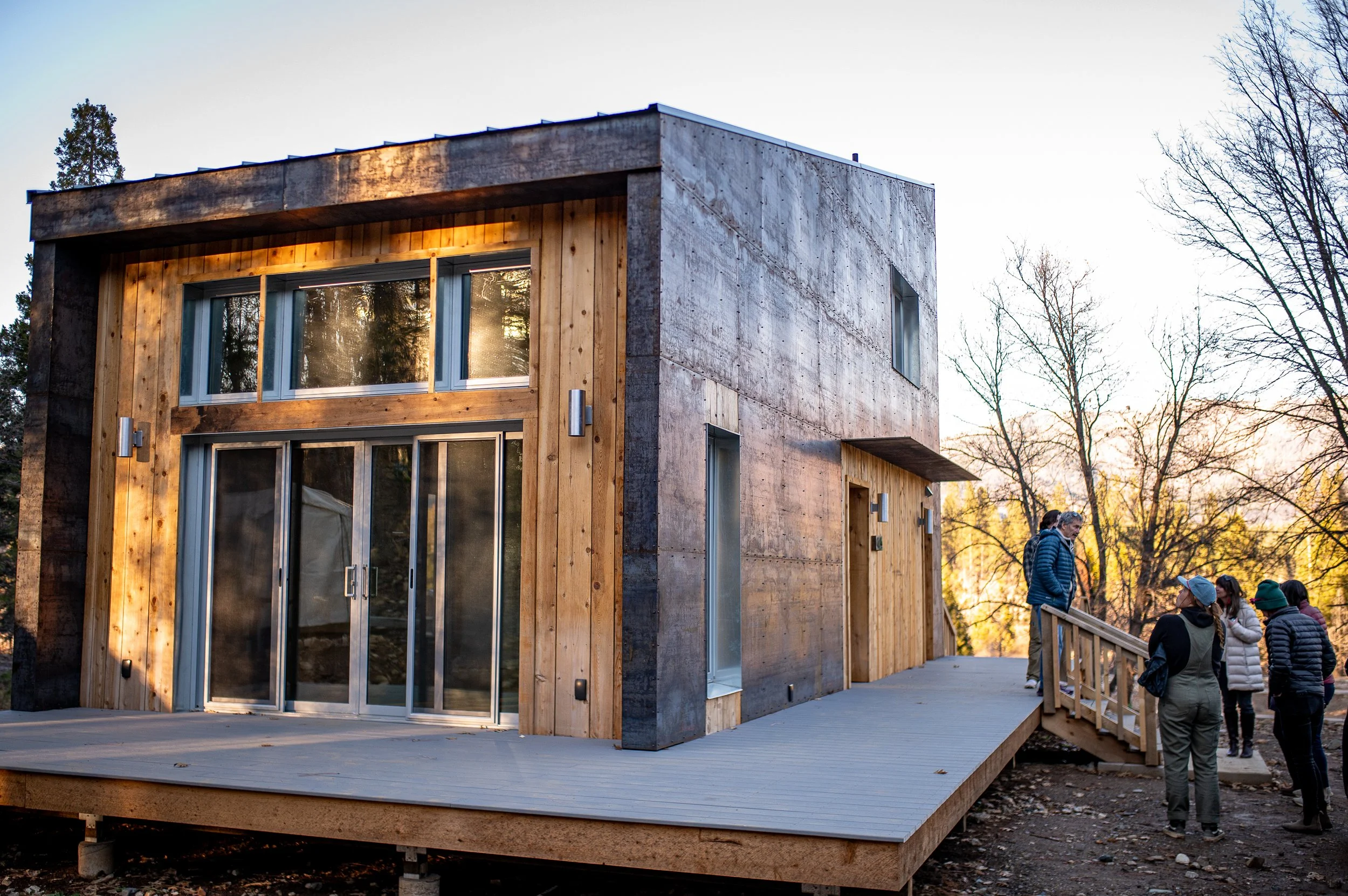A modern two-story CLT home with a Mass timber wooden and steel exterior, large sliding glass doors, and a spacious deck, surrounded by trees with some autumn foliage, with a group of people standing nearby.