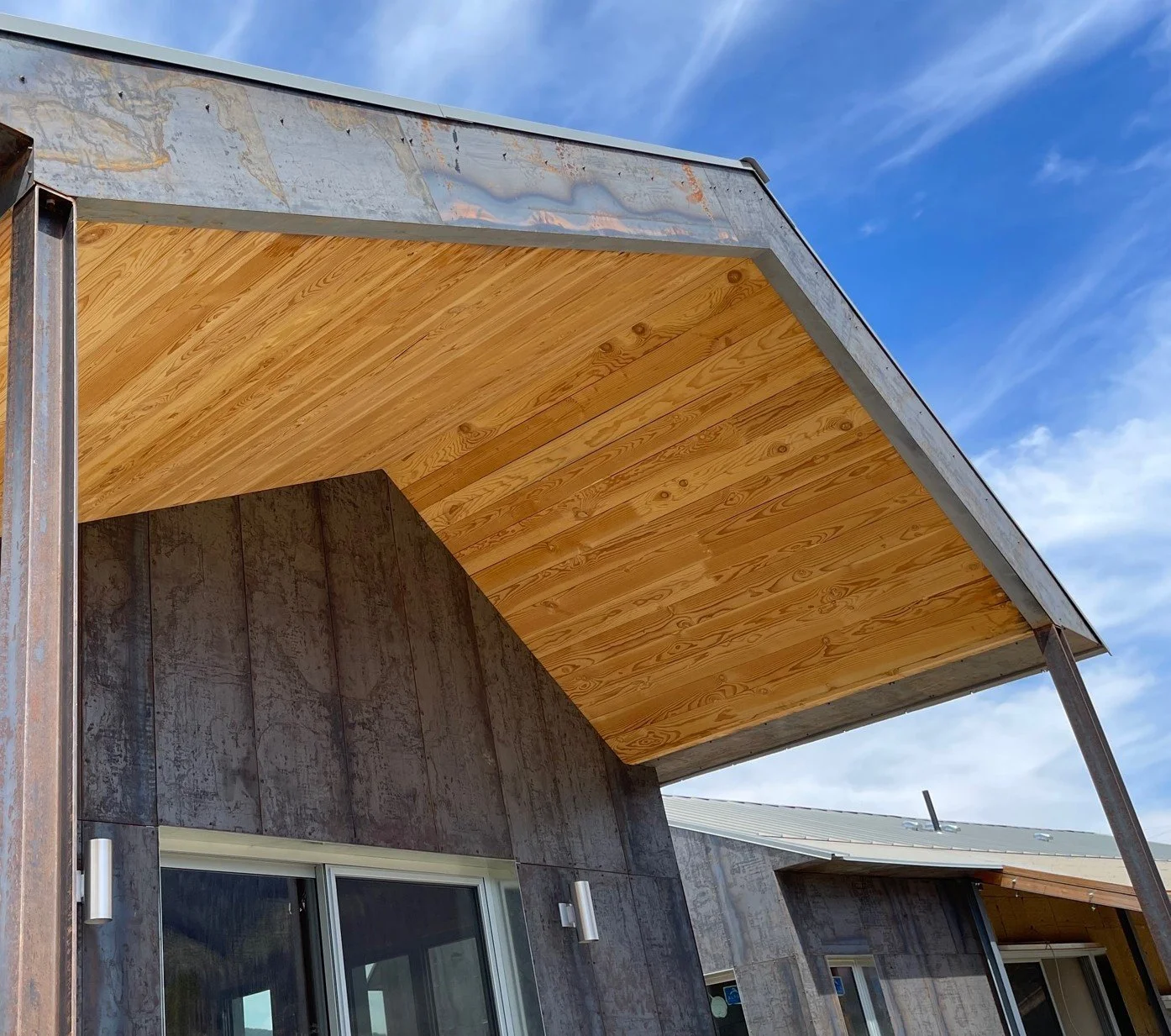 Mass Timber home exterior with a steel panel exterior, wooden underside exposed on the porch roof, and blue sky with clouds.