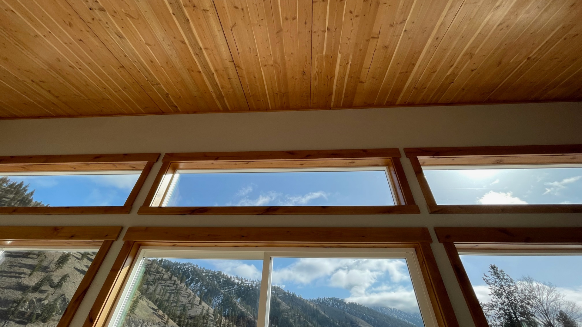 Interior of a room with large, angled windows showing a mountain landscape and blue sky with clouds outside, wooden CLT ceiling, and trim around the windows.
