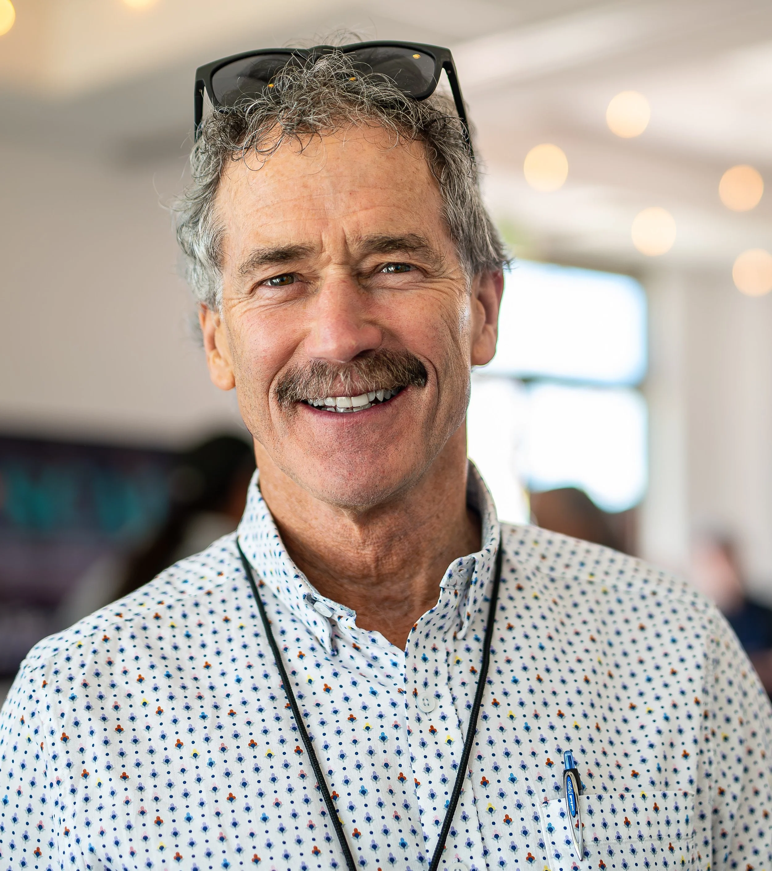 Jonathan Kusel, Executive Director, A smiling middle-aged man with gray hair, a mustache, and glasses resting on his head, wearing a white shirt with colored polka dots, in a bright indoor setting.